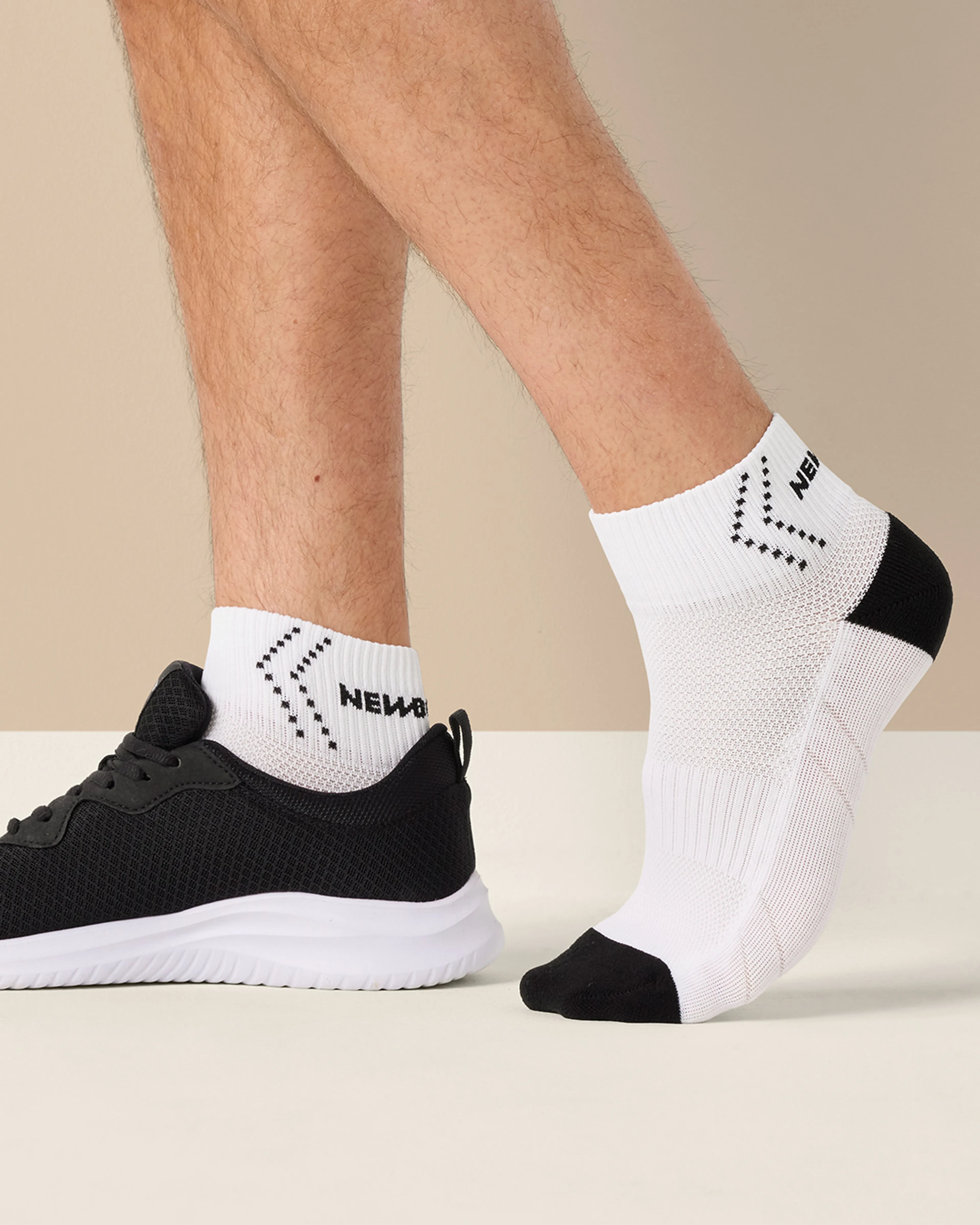 Ett par vita strumpor med svart tå- och hälparti, designade för prestation och komfort, visas på en mans fot bredvid en svart sportig sko. Strumporna har också svart mönster och text vid skaftet.