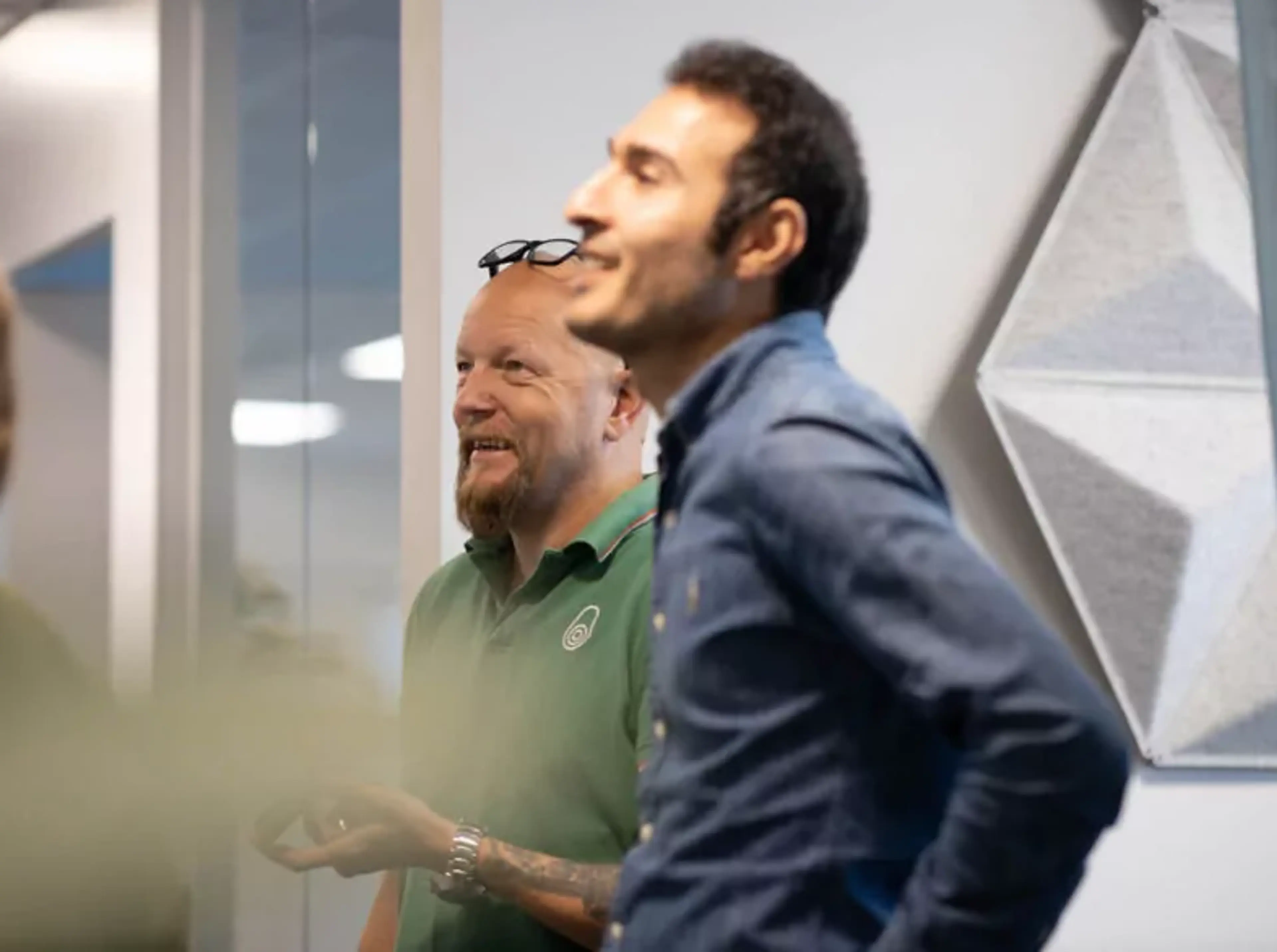 Tre personer i en kontorsmiljö mitt i ett samtal, med ljusa väggar och en geometrisk dekorpanel i bakgrunden.