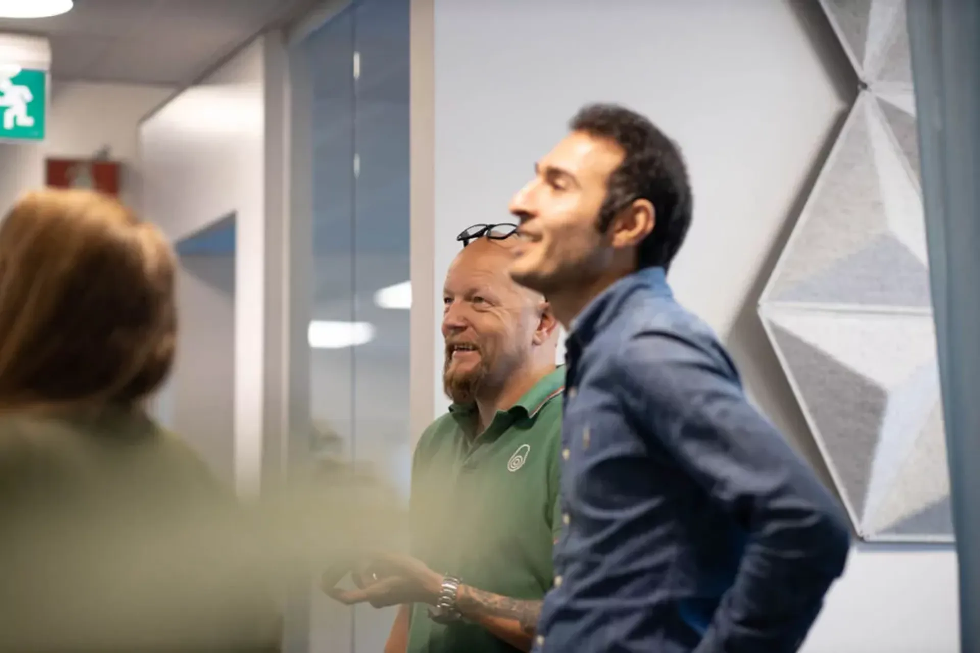 Tre personer i en kontorsmiljö mitt i ett samtal, med ljusa väggar och en geometrisk dekorpanel i bakgrunden.