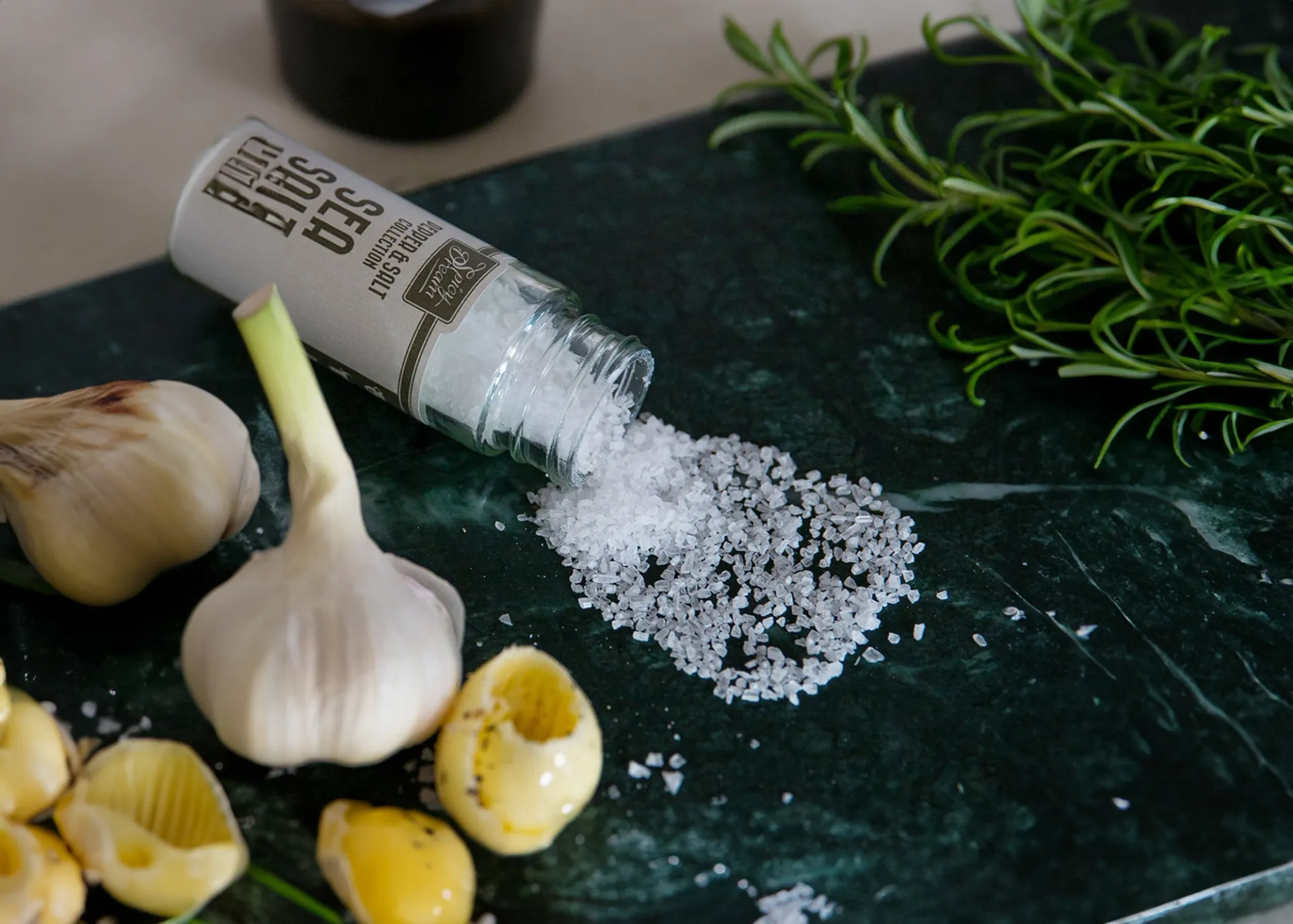 En glasburk med grovt havssalt som är urtappat på en mörk grön marmorbänk. I bakgrunden syns en vitlök, vitlöksskivor och en kvist rosmarin.