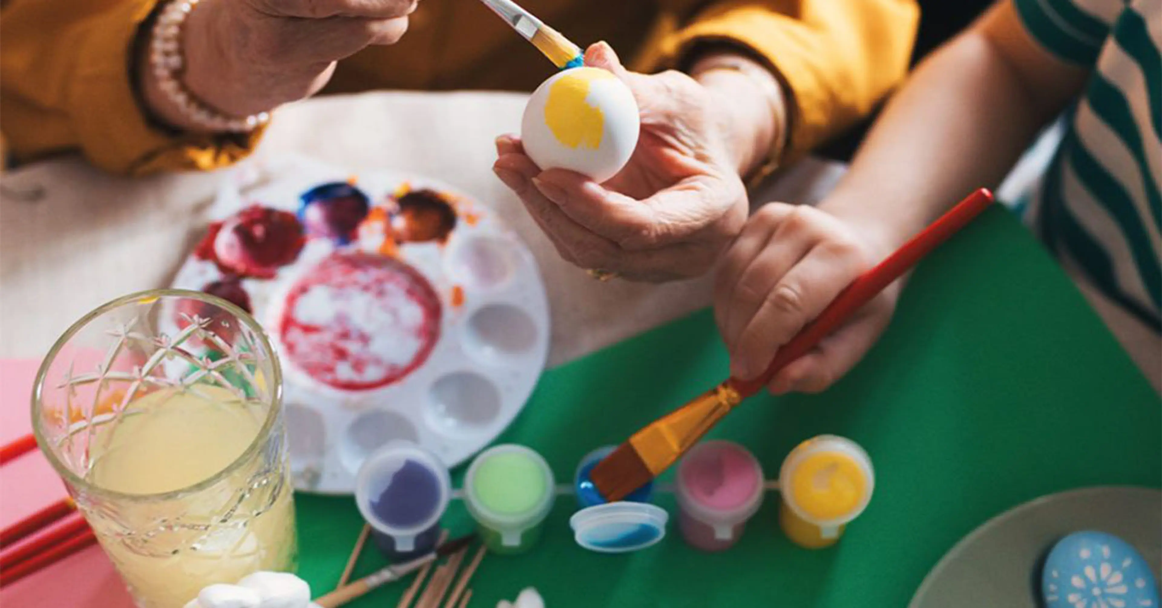 Två personer målar påskägg med färgglada penseldrag, omgivna av små keramikharar och flera färgburkar. Ett fat med färdigmålade ägg och en mugg med ljus dryck står också på bordet.