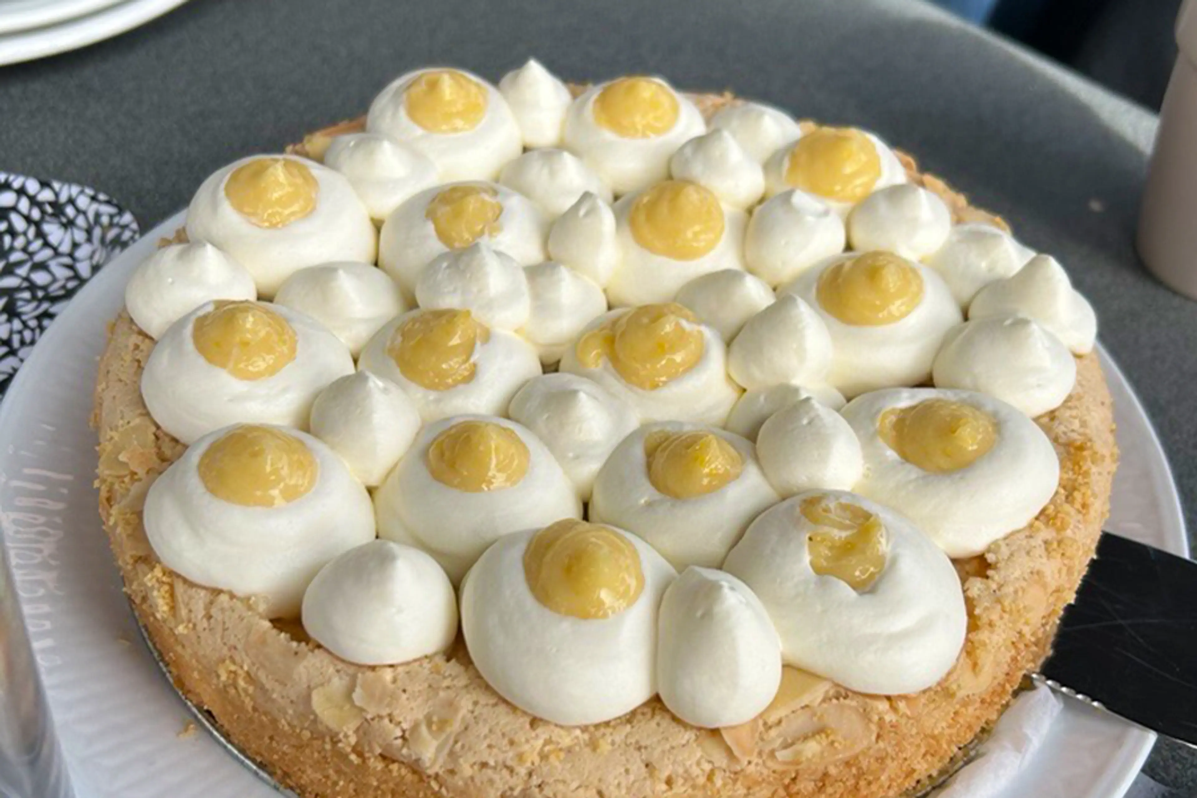 En påsktårta med krispig botten, toppad med regelbundet spritsade klickar av vit grädde och gula citronsåser. Tårtan står på ett vitt fat på ett mörkt bord.