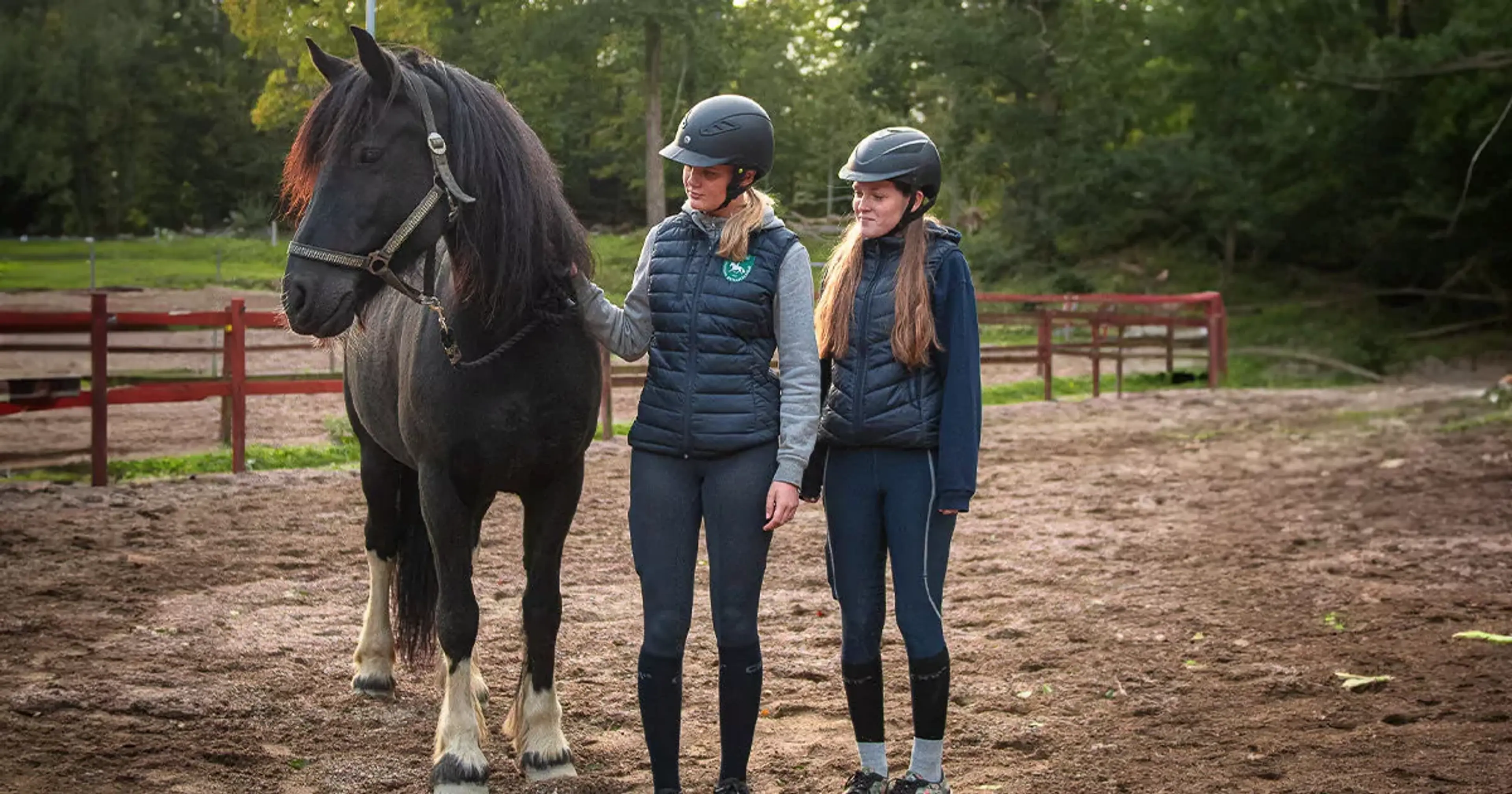 Två tjejer i ridkläder och hjälmar står bredvid en svart häst med vita strumpor på bakbenen på en ridbana omgiven av grönska. De ser lugna ut och håller hästen i tyglarna.