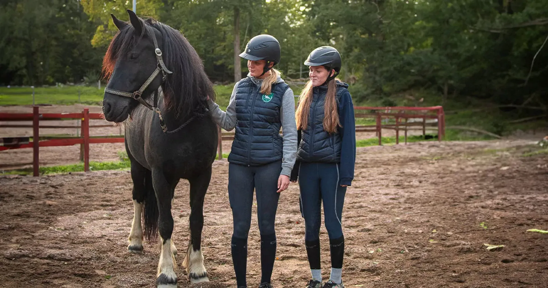 Två tjejer i ridkläder och hjälmar står bredvid en svart häst med vita strumpor på bakbenen på en ridbana omgiven av grönska. De ser lugna ut och håller hästen i tyglarna.