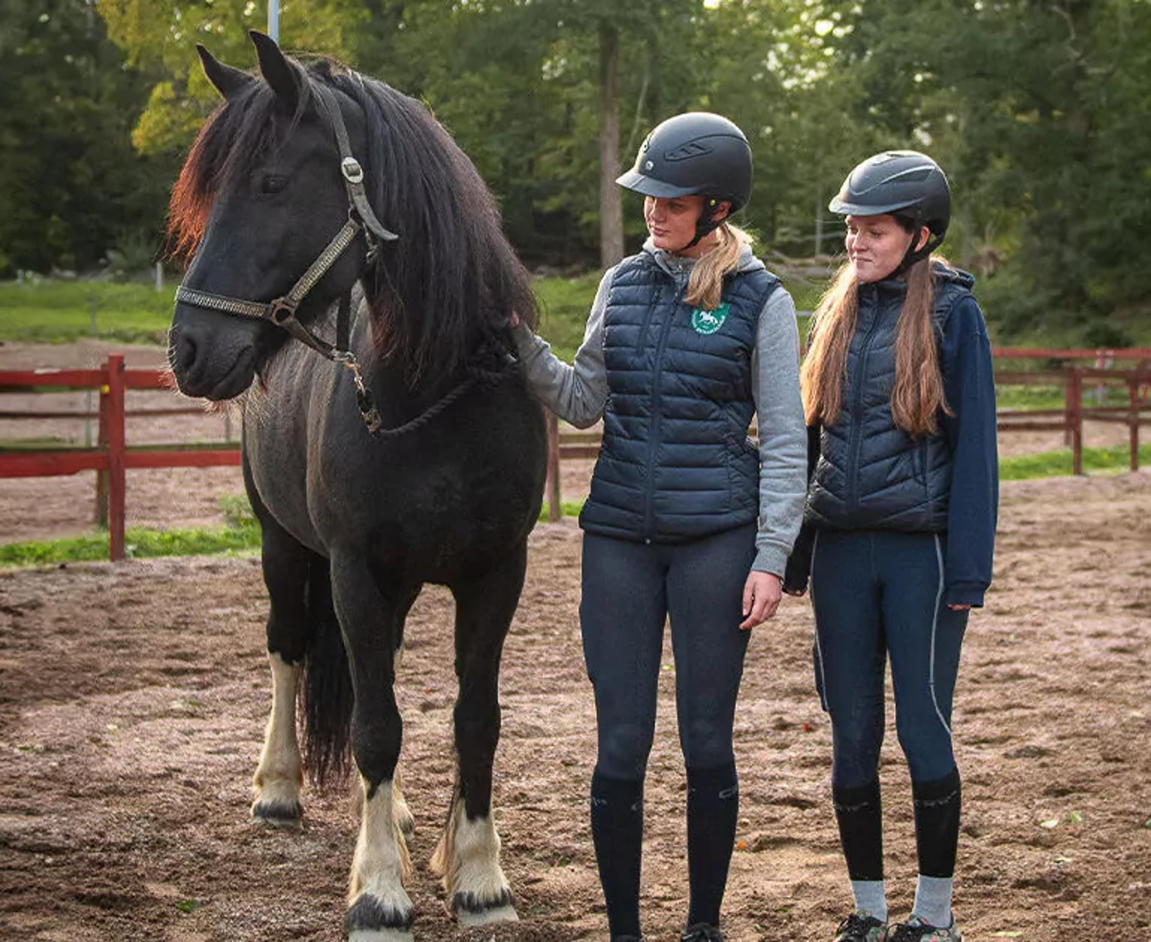 Två tjejer i ridkläder och hjälmar står bredvid en svart häst med vita strumpor på bakbenen på en ridbana omgiven av grönska. De ser lugna ut och håller hästen i tyglarna.