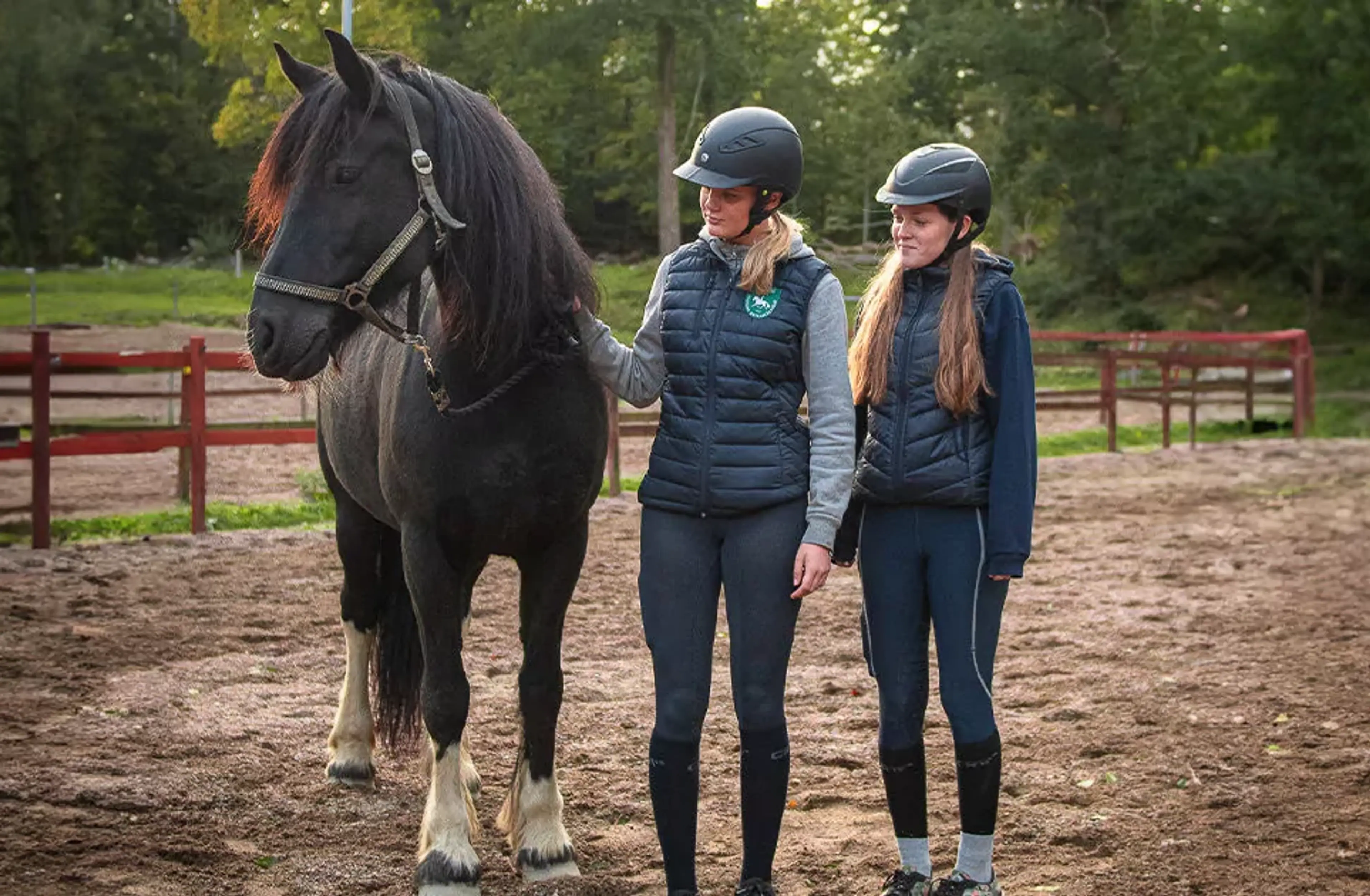 Två tjejer i ridkläder och hjälmar står bredvid en svart häst med vita strumpor på bakbenen på en ridbana omgiven av grönska. De ser lugna ut och håller hästen i tyglarna.