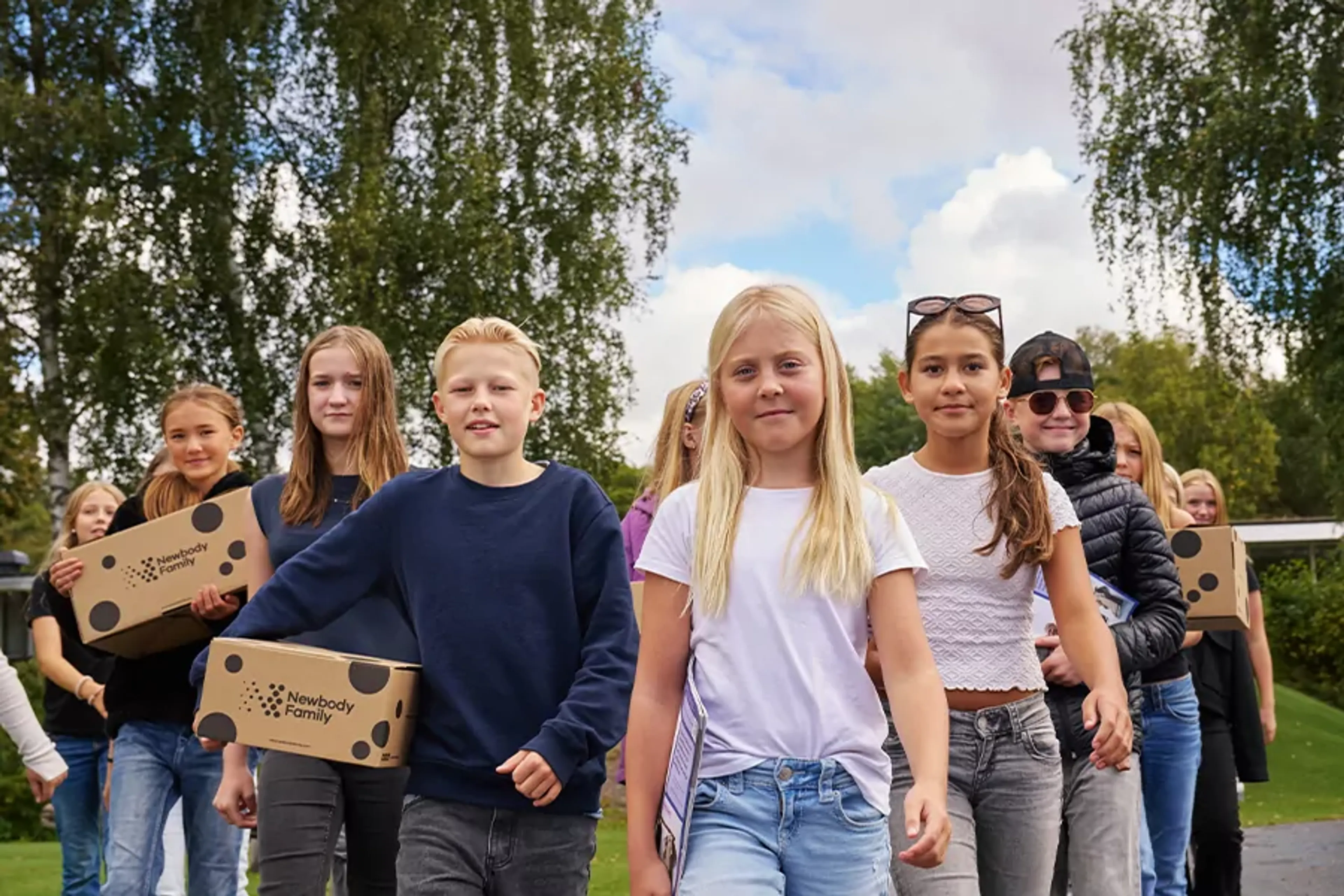 En grupp barn går tillsammans utomhus med kartonger i händerna, omgivna av grönska och blå himmel. De ser ut att vara på väg någonstans, fulla av energi och samarbete.
