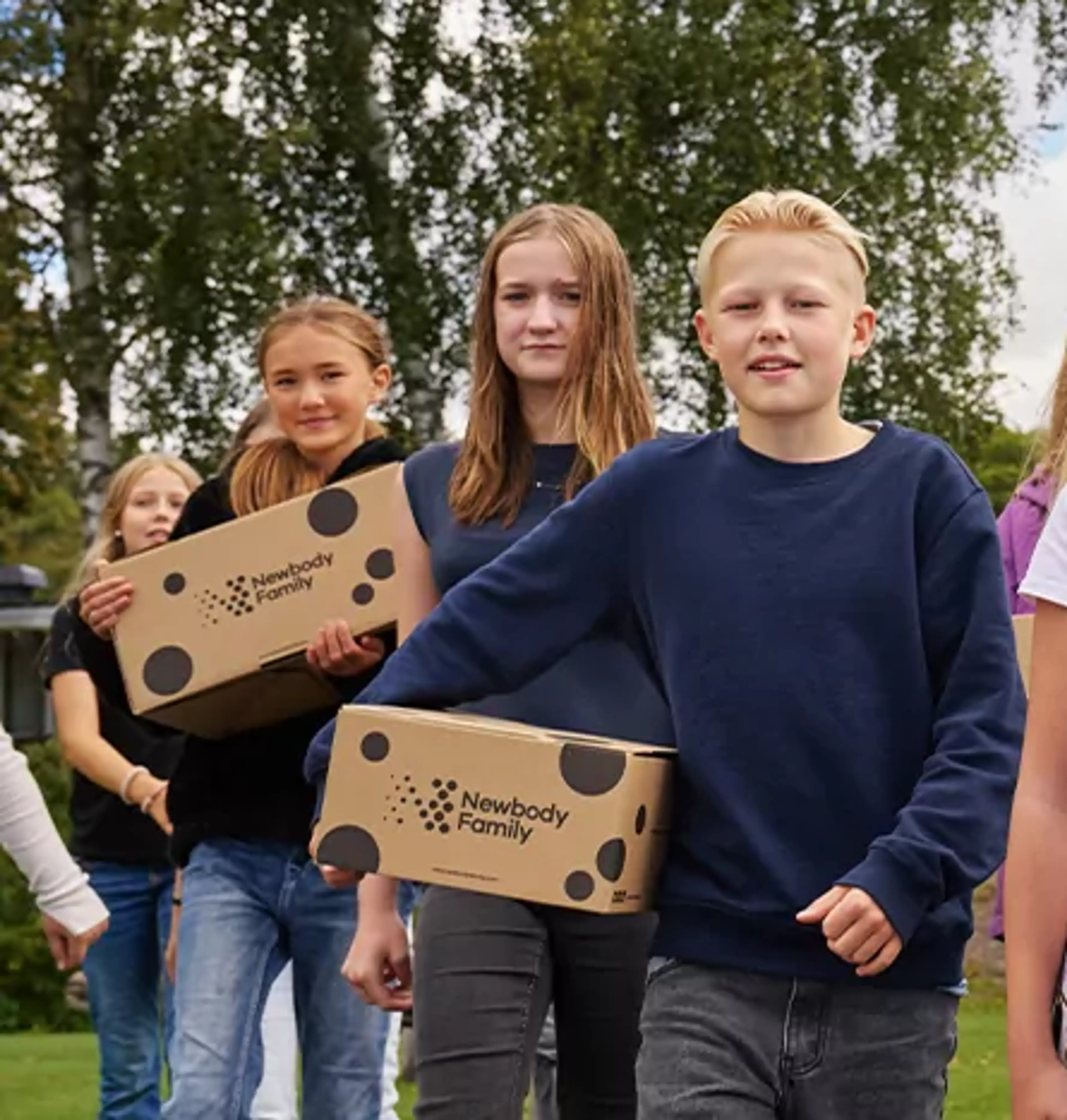 En grupp barn går tillsammans utomhus med kartonger i händerna, omgivna av grönska och blå himmel. De ser ut att vara på väg någonstans, fulla av energi och samarbete.