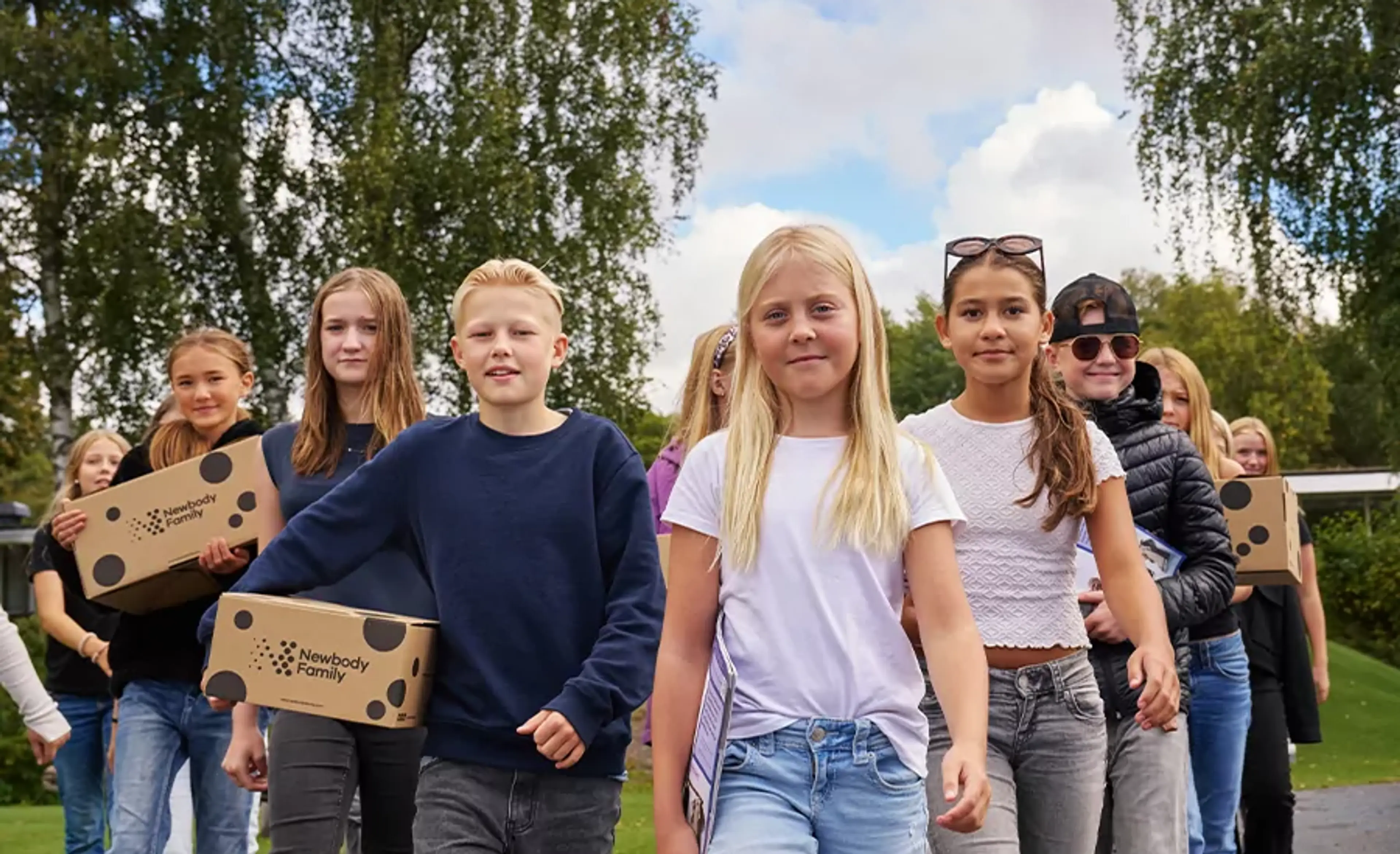En grupp barn går tillsammans utomhus med kartonger i händerna, omgivna av grönska och blå himmel. De ser ut att vara på väg någonstans, fulla av energi och samarbete.