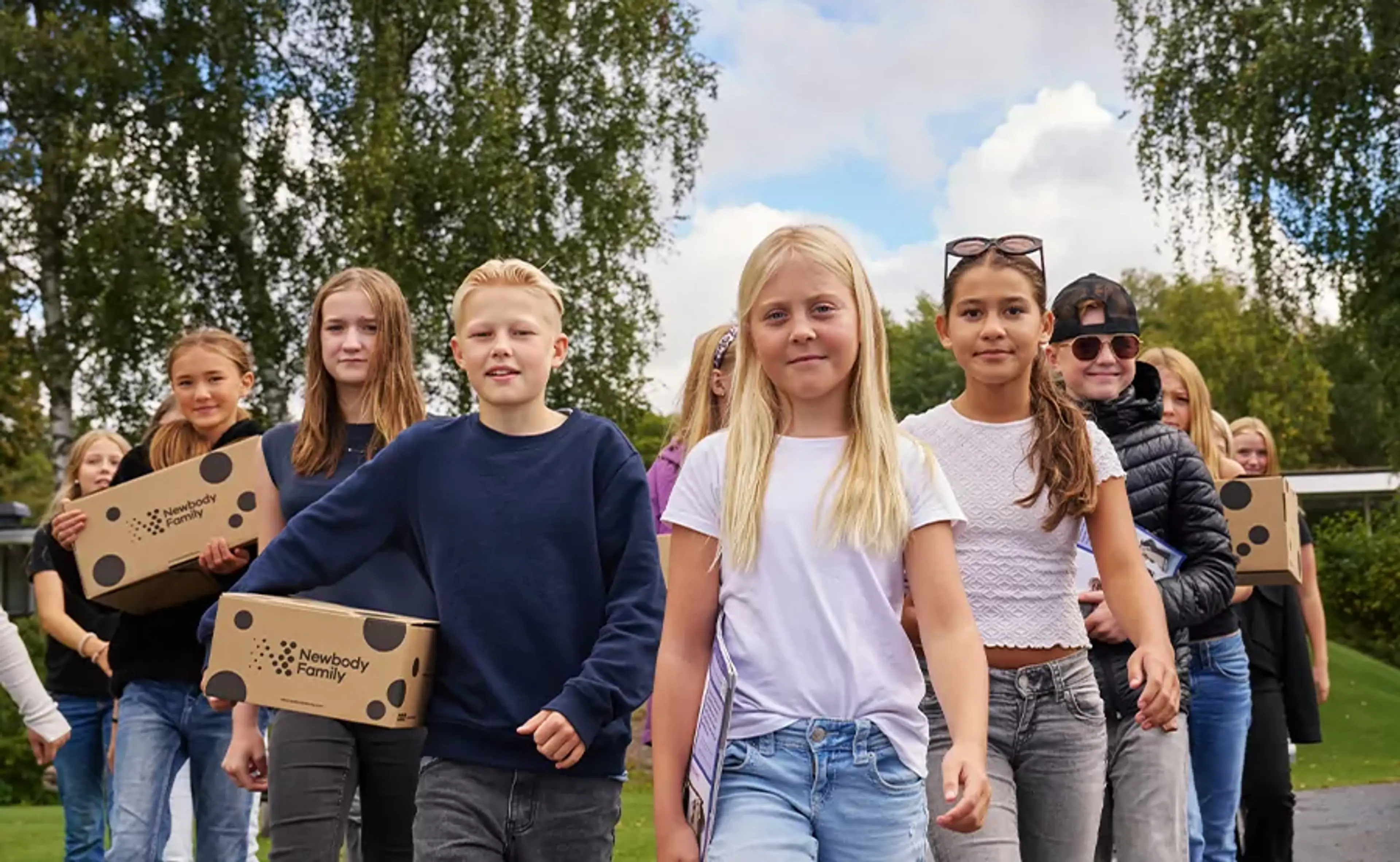 En grupp barn går tillsammans utomhus med kartonger i händerna, omgivna av grönska och blå himmel. De ser ut att vara på väg någonstans, fulla av energi och samarbete.