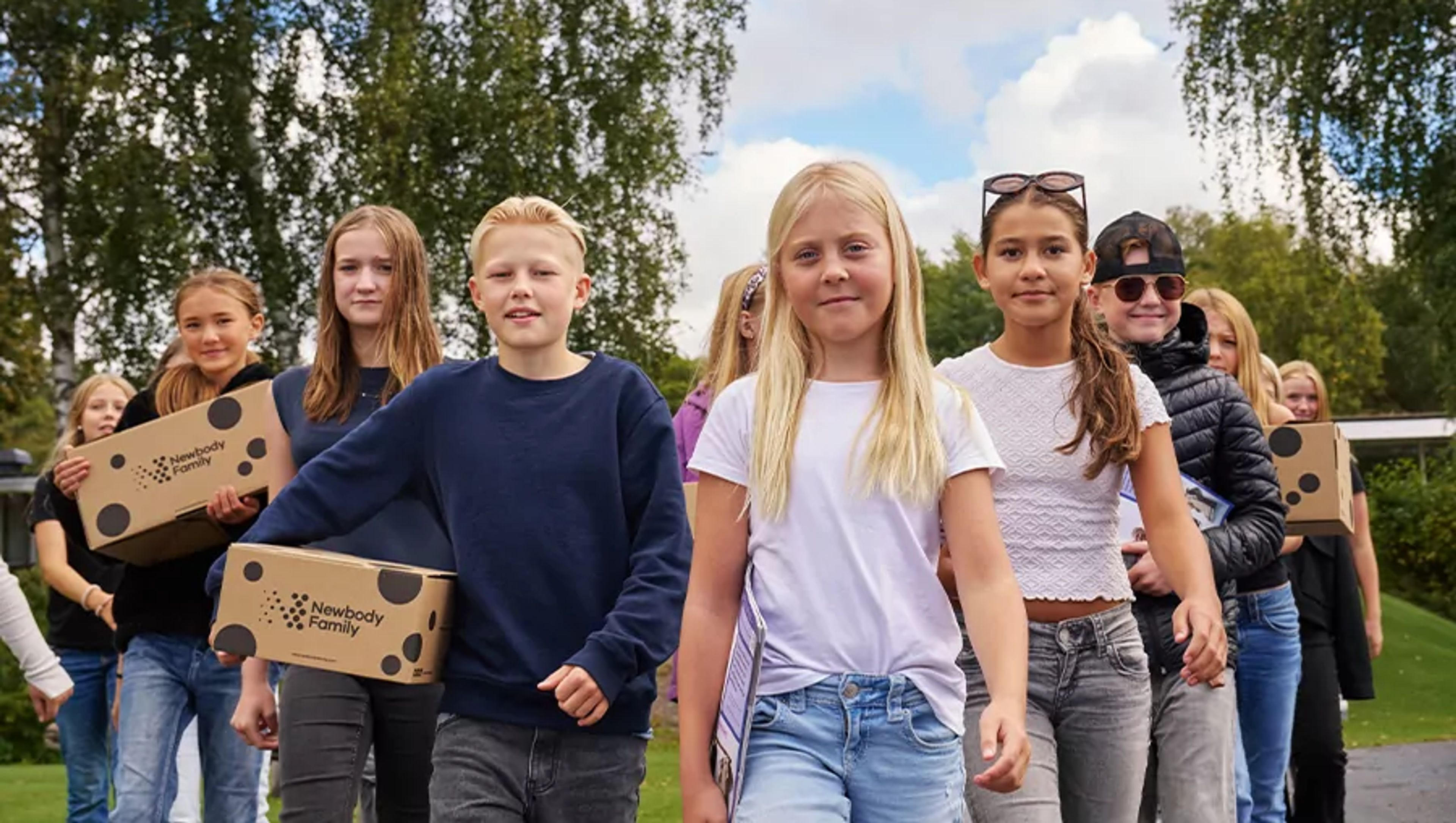 En grupp barn går tillsammans utomhus med kartonger i händerna, omgivna av grönska och blå himmel. De ser ut att vara på väg någonstans, fulla av energi och samarbete.