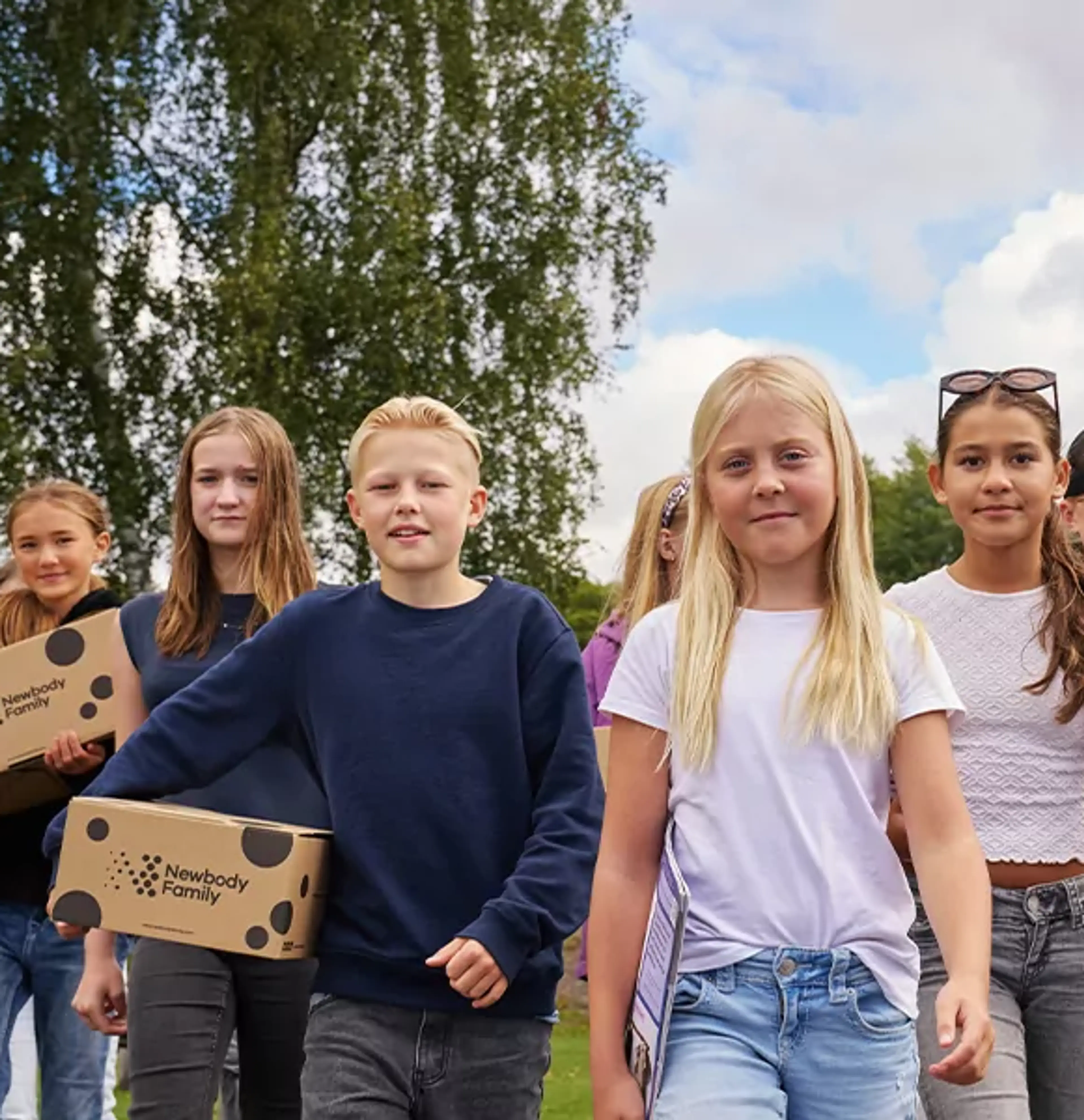 En grupp barn går tillsammans utomhus med kartonger i händerna, omgivna av grönska och blå himmel. De ser ut att vara på väg någonstans, fulla av energi och samarbete.