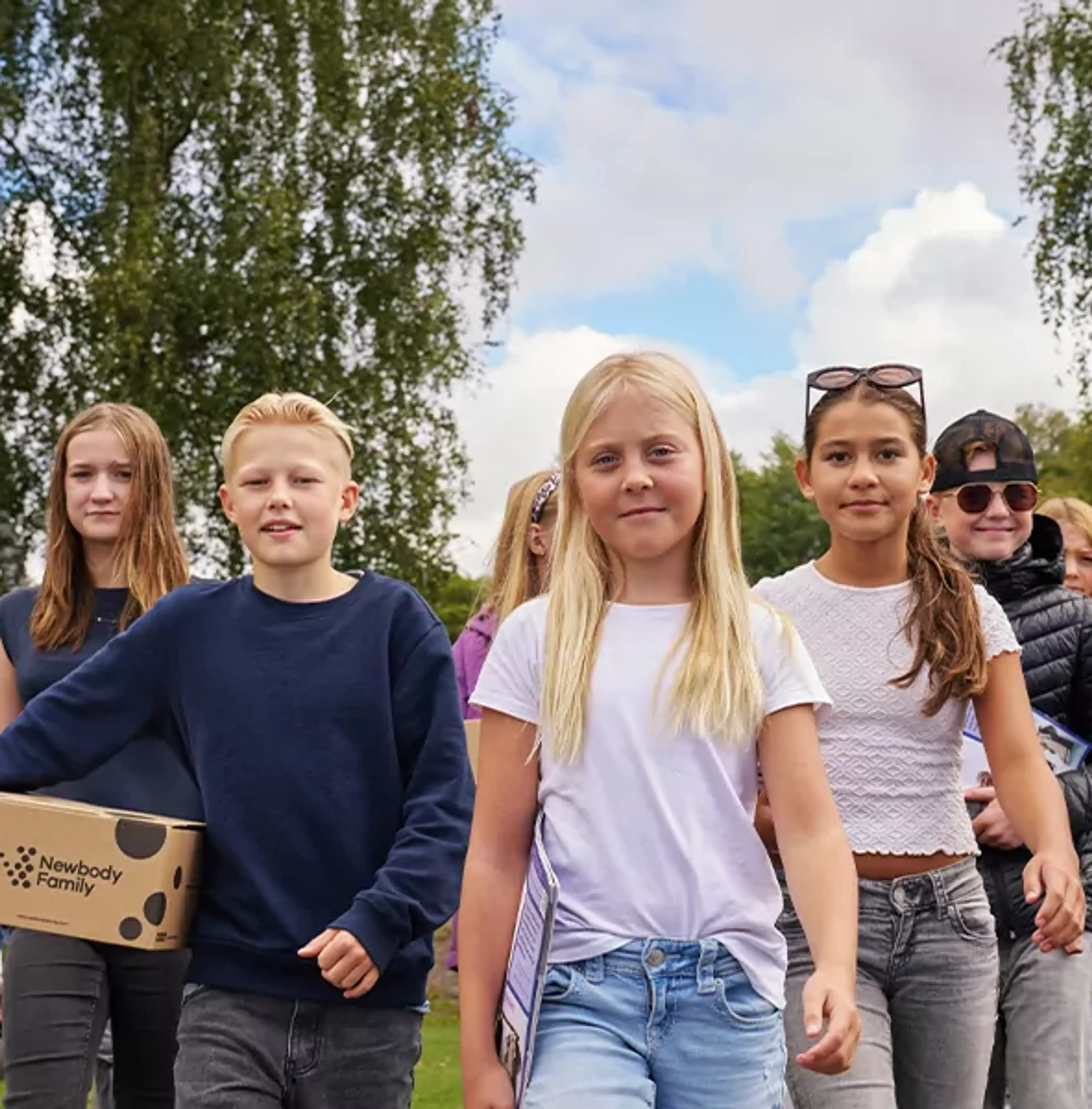En grupp barn går tillsammans utomhus med kartonger i händerna, omgivna av grönska och blå himmel. De ser ut att vara på väg någonstans, fulla av energi och samarbete.