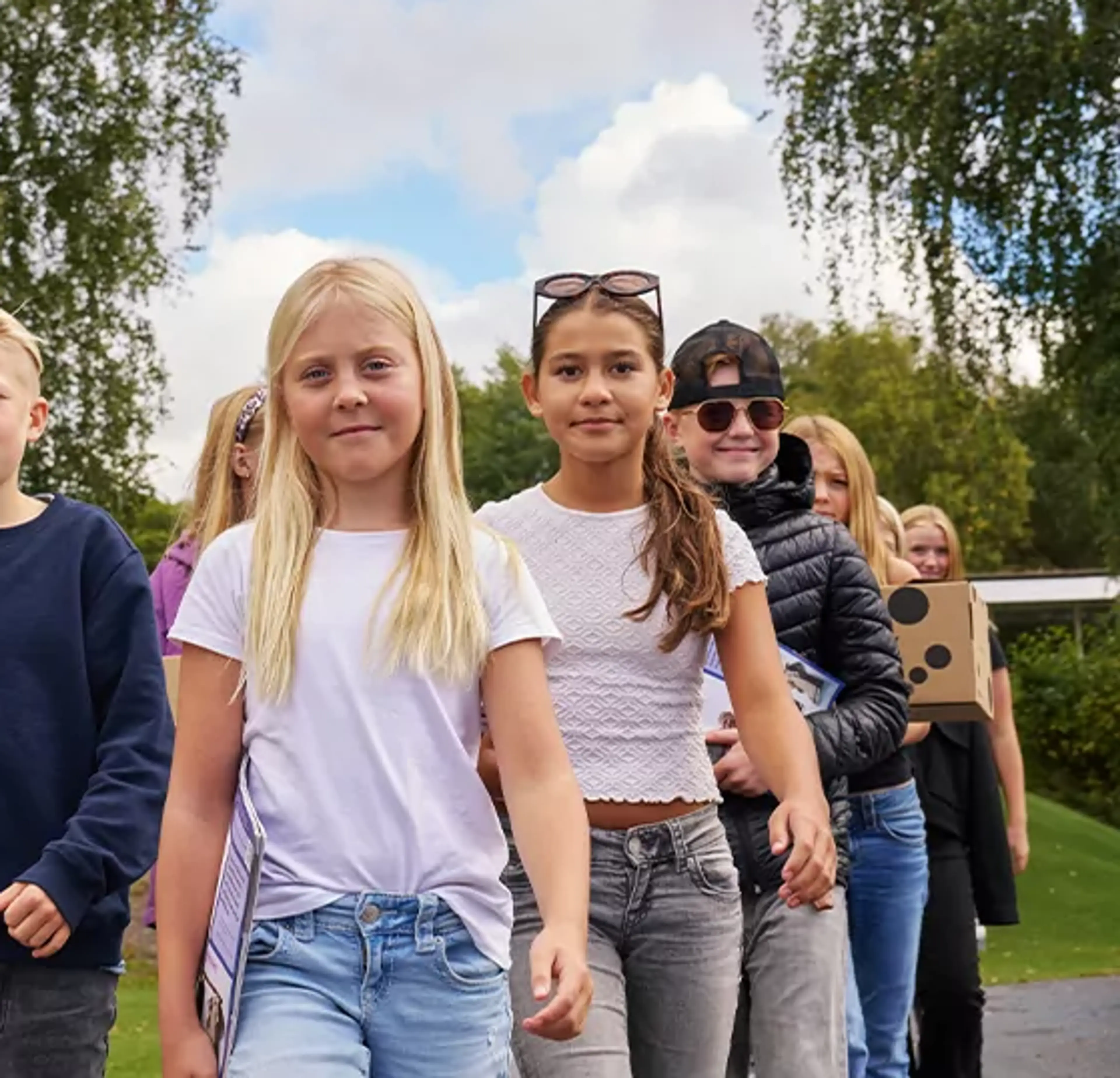 En grupp barn går tillsammans utomhus med kartonger i händerna, omgivna av grönska och blå himmel. De ser ut att vara på väg någonstans, fulla av energi och samarbete.