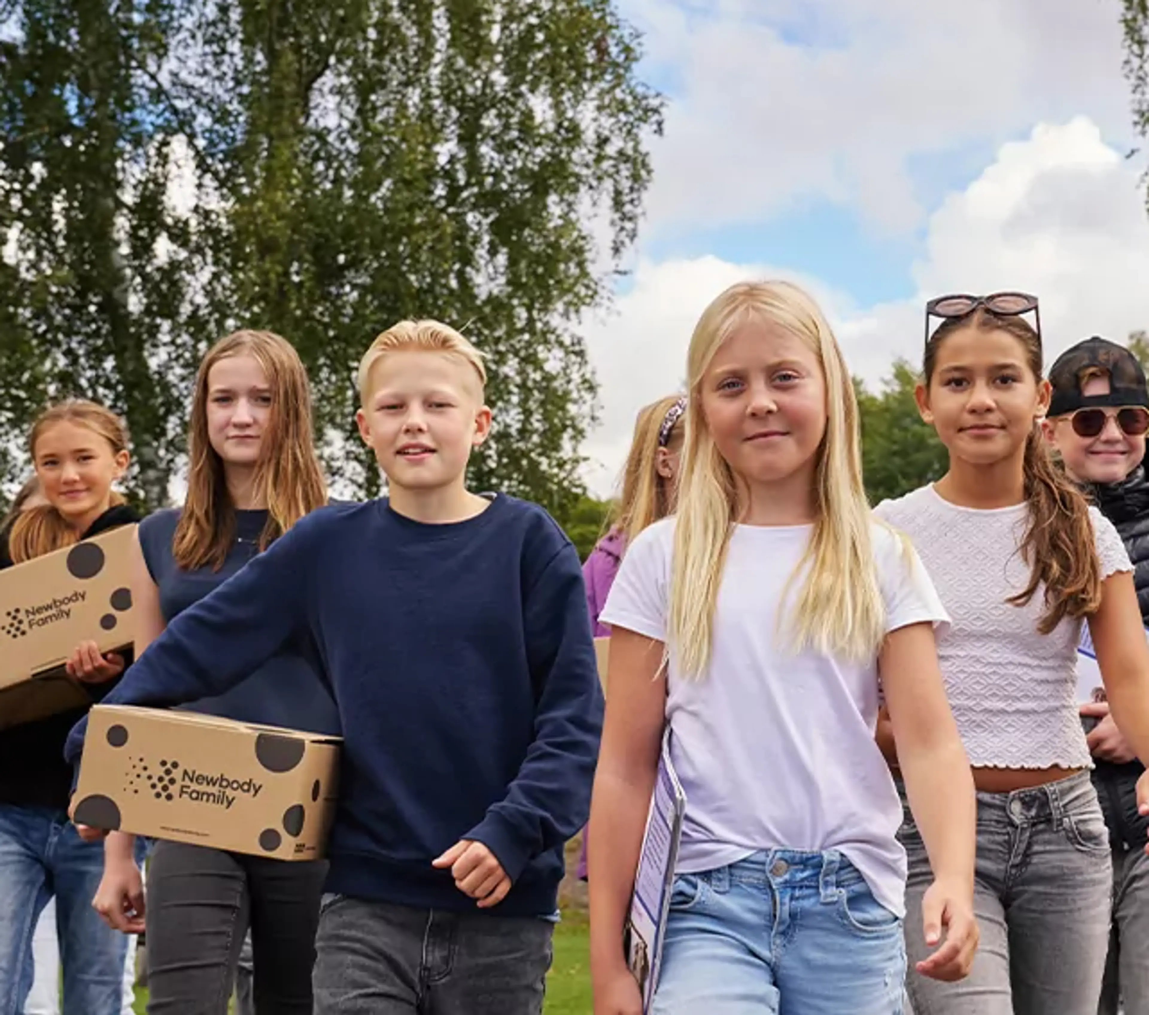 En grupp barn går tillsammans utomhus med kartonger i händerna, omgivna av grönska och blå himmel. De ser ut att vara på väg någonstans, fulla av energi och samarbete.