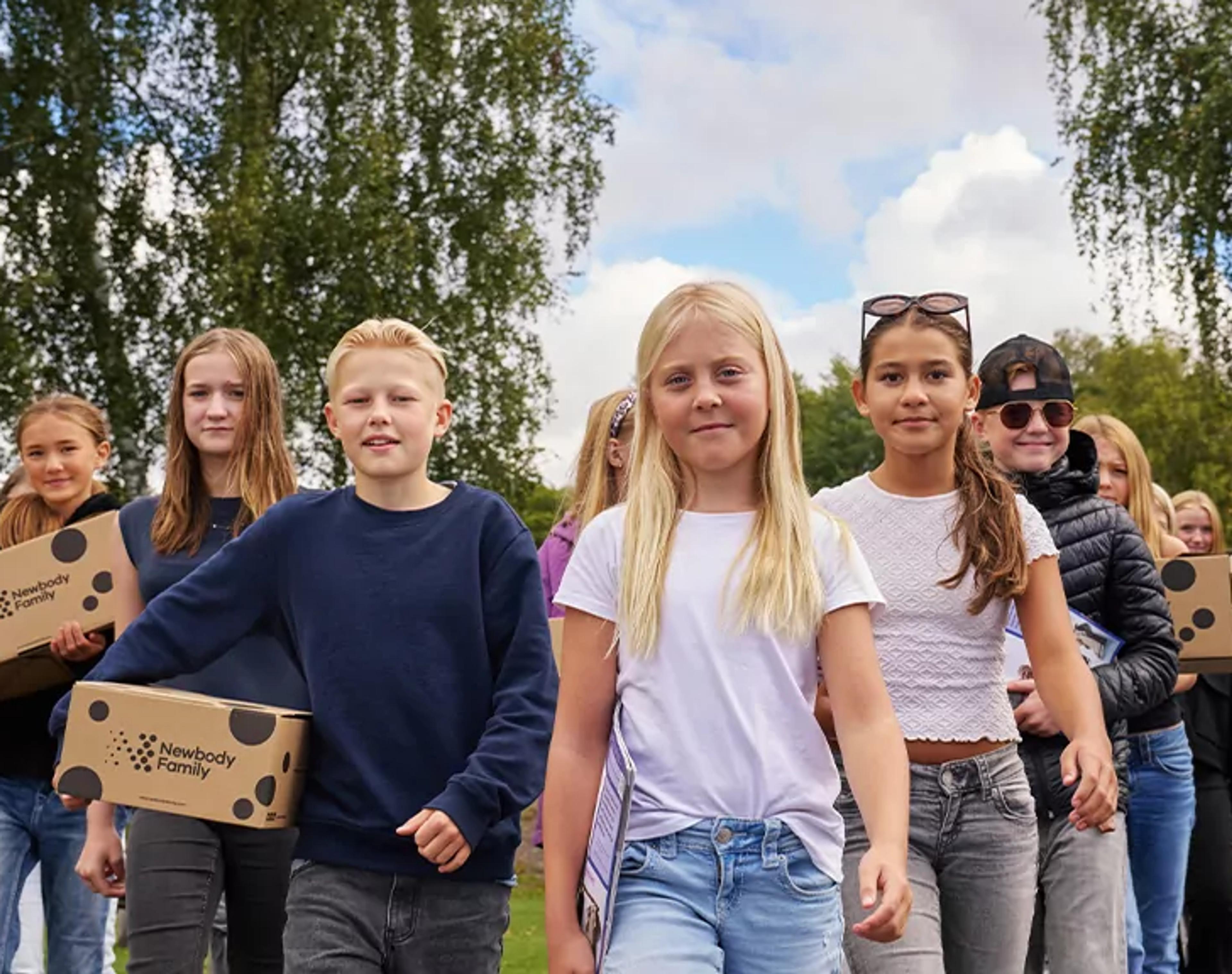 En grupp barn går tillsammans utomhus med kartonger i händerna, omgivna av grönska och blå himmel. De ser ut att vara på väg någonstans, fulla av energi och samarbete.