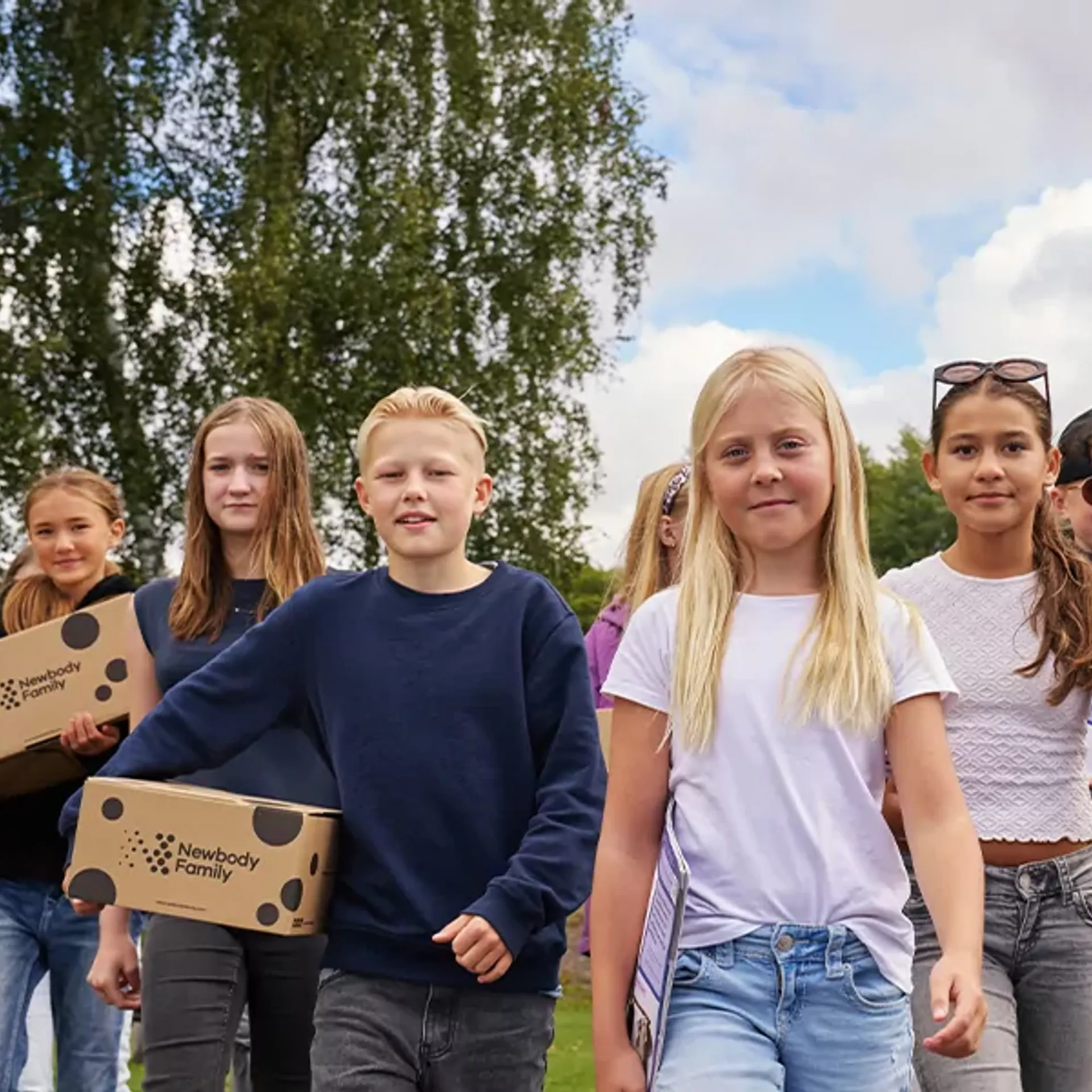 En grupp barn går tillsammans utomhus med kartonger i händerna, omgivna av grönska och blå himmel. De ser ut att vara på väg någonstans, fulla av energi och samarbete.