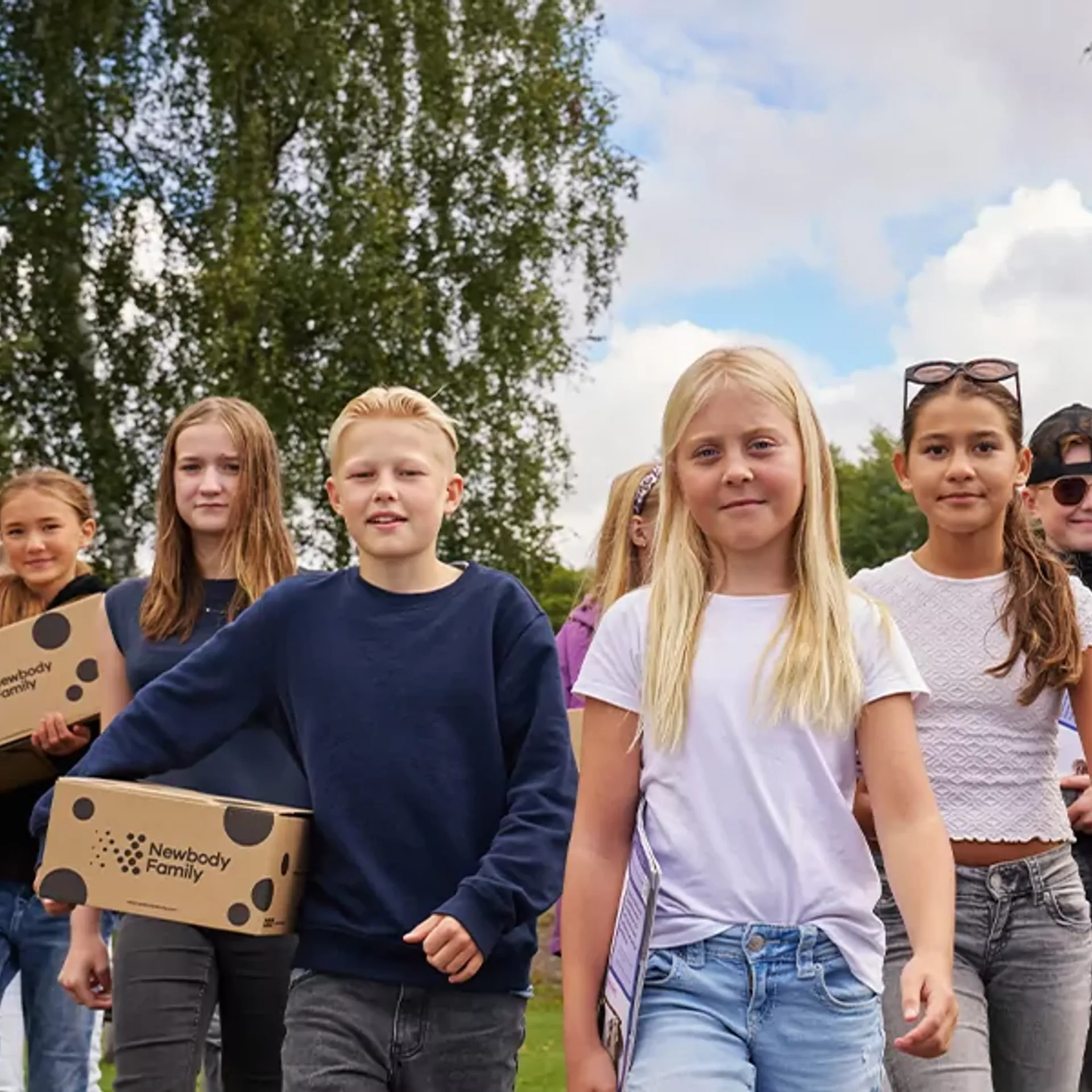 En grupp barn går tillsammans utomhus med kartonger i händerna, omgivna av grönska och blå himmel. De ser ut att vara på väg någonstans, fulla av energi och samarbete.