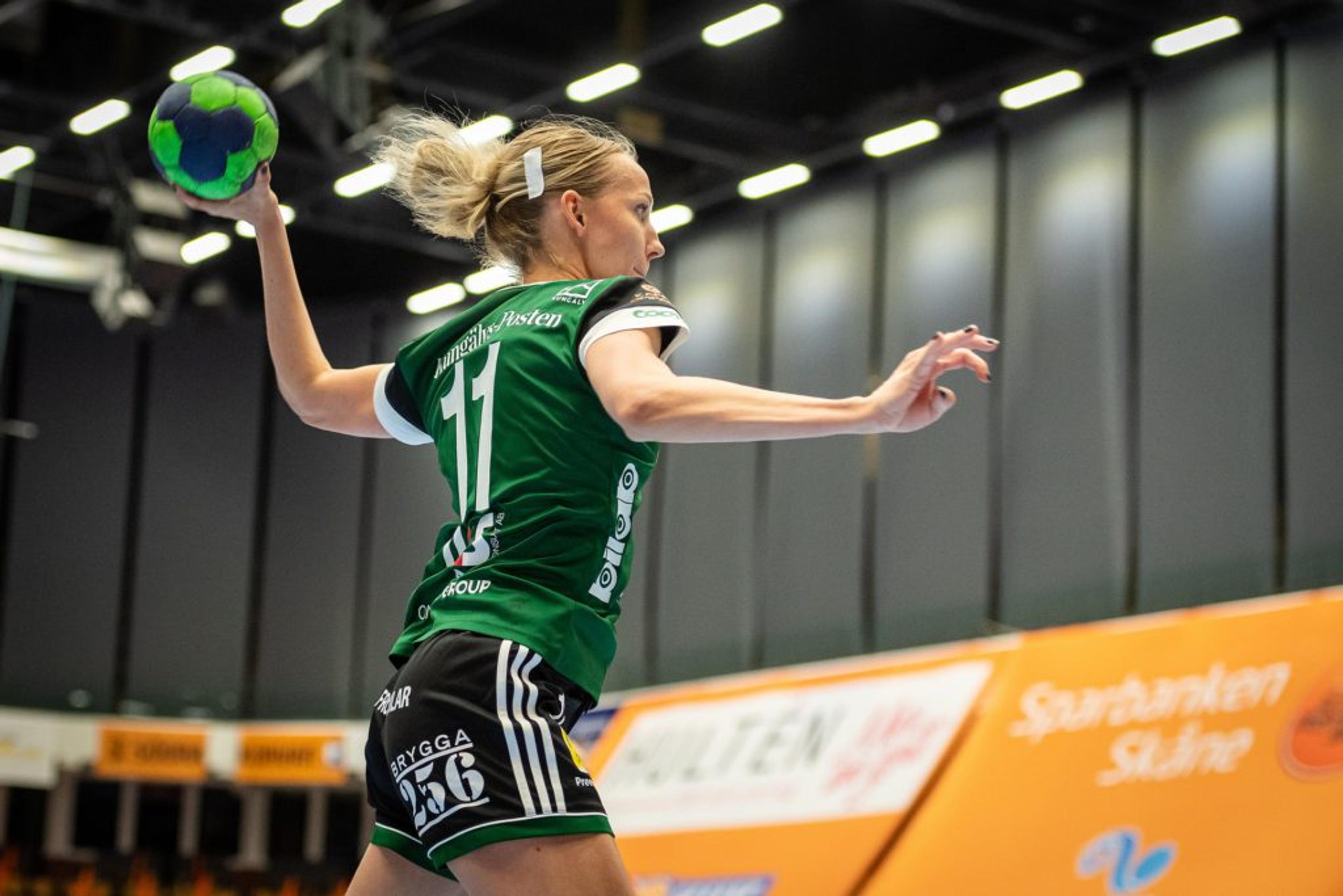 En handbollsspelare i grön tröja hoppar för att kasta en blå-grön boll i en inomhushall. Bakgrunden visar reklamskyltar och högt i tak.