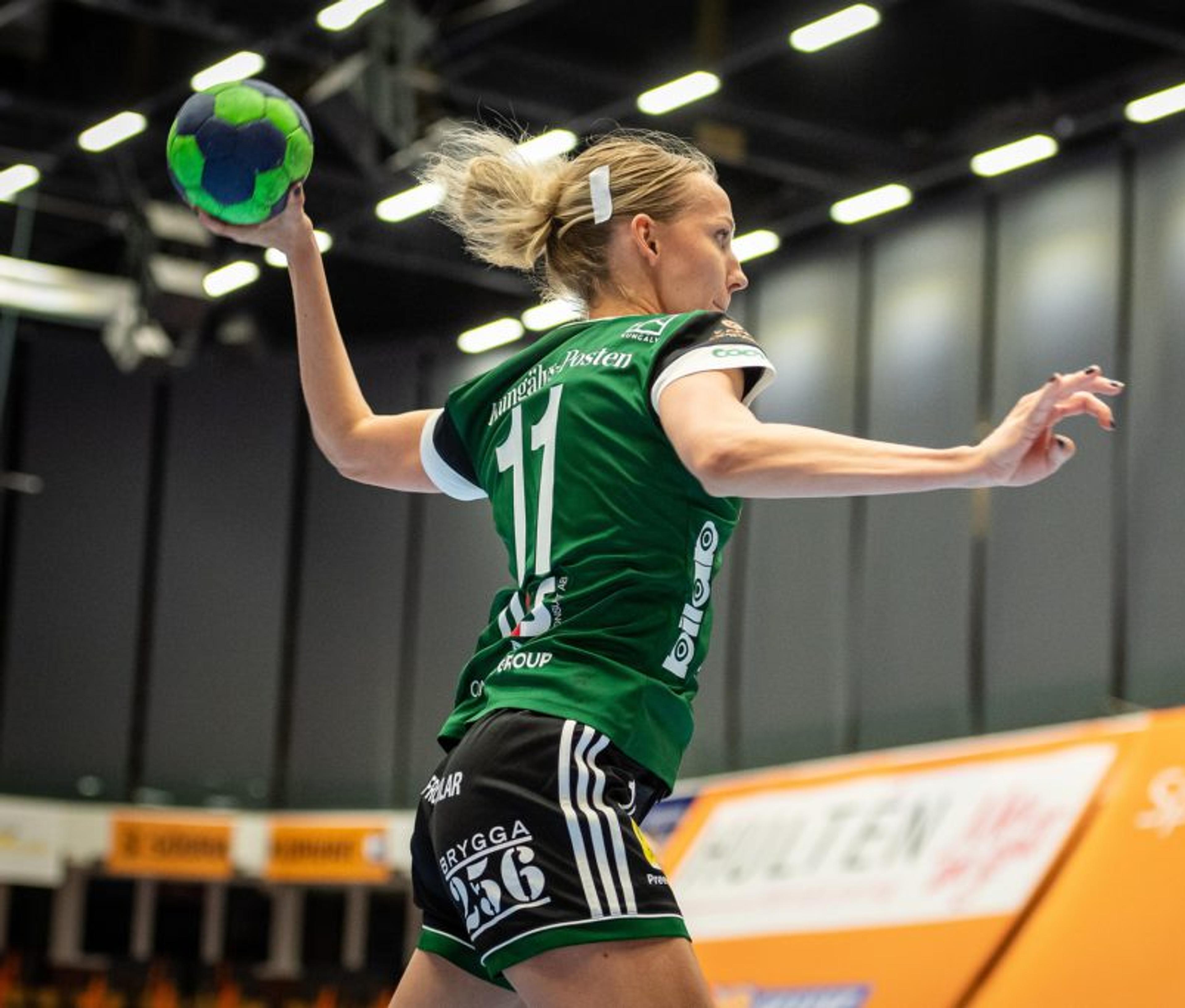En handbollsspelare i grön tröja hoppar för att kasta en blå-grön boll i en inomhushall. Bakgrunden visar reklamskyltar och högt i tak.