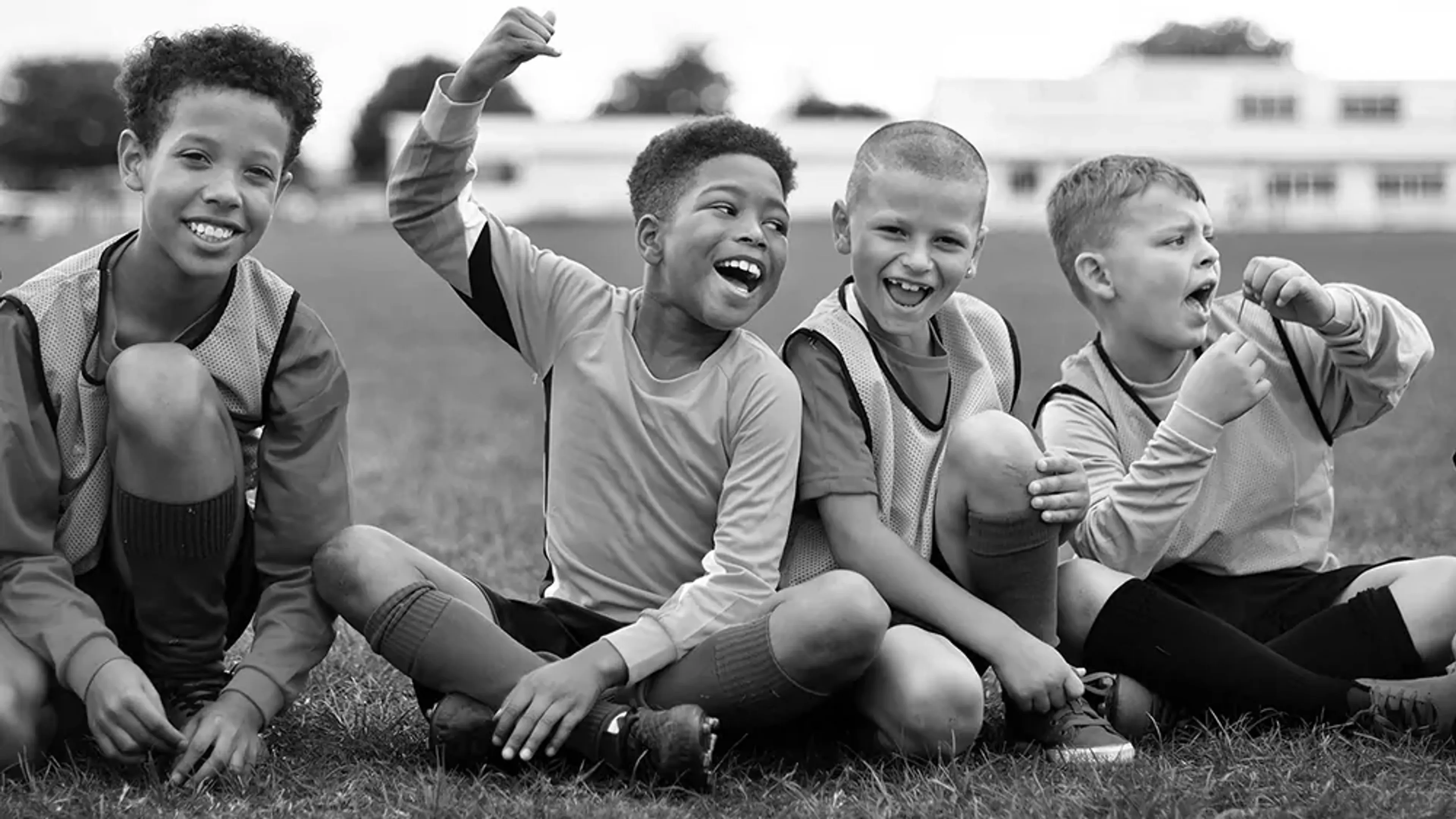 Fyra barn sitter på gräset i fotbollskläder och skrattar tillsammans efter en match eller träning. De visar glädje och samhörighet i en lekfull stund utomhus.