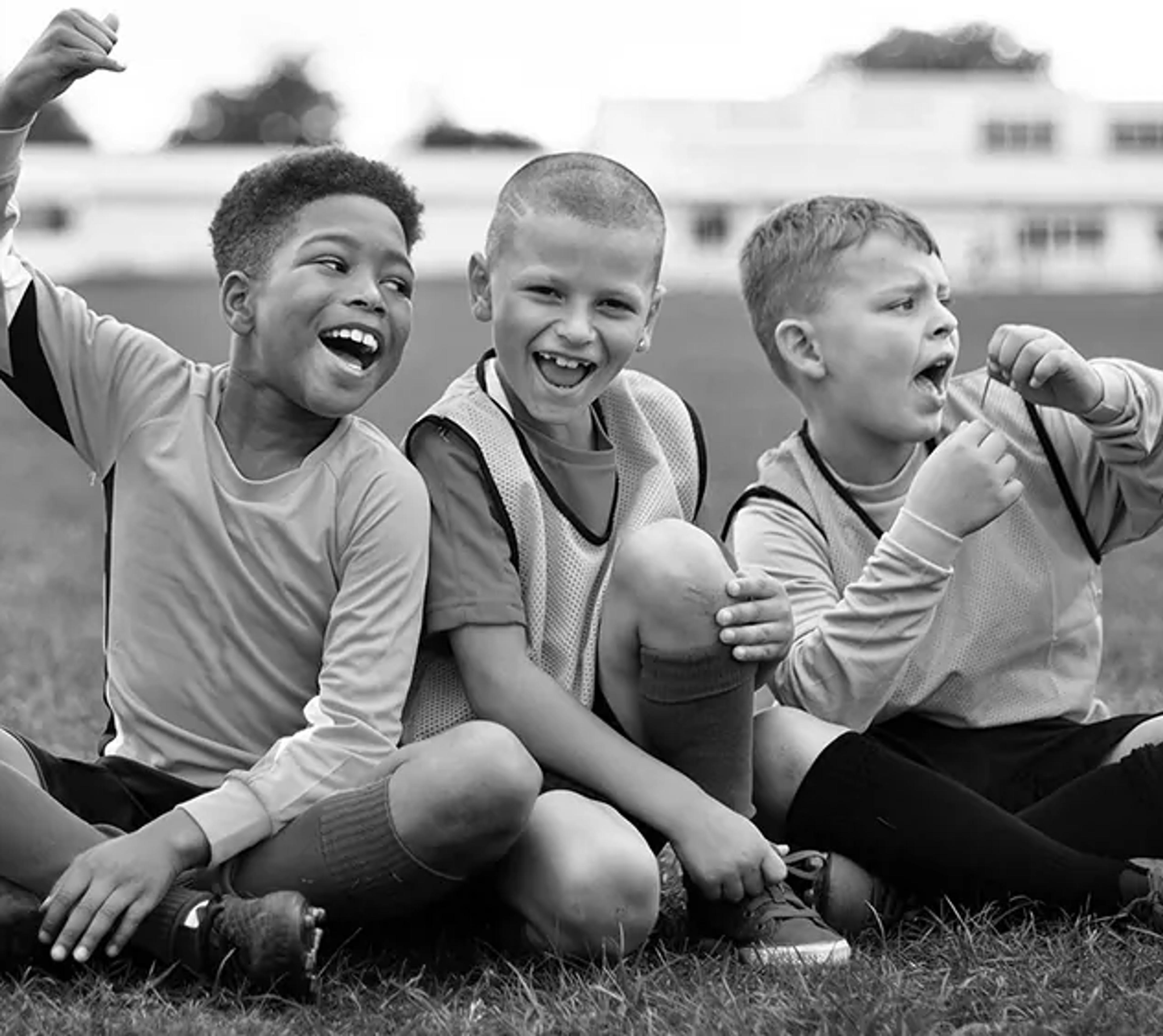 Fyra barn sitter på gräset i fotbollskläder och skrattar tillsammans efter en match eller träning. De visar glädje och samhörighet i en lekfull stund utomhus.
