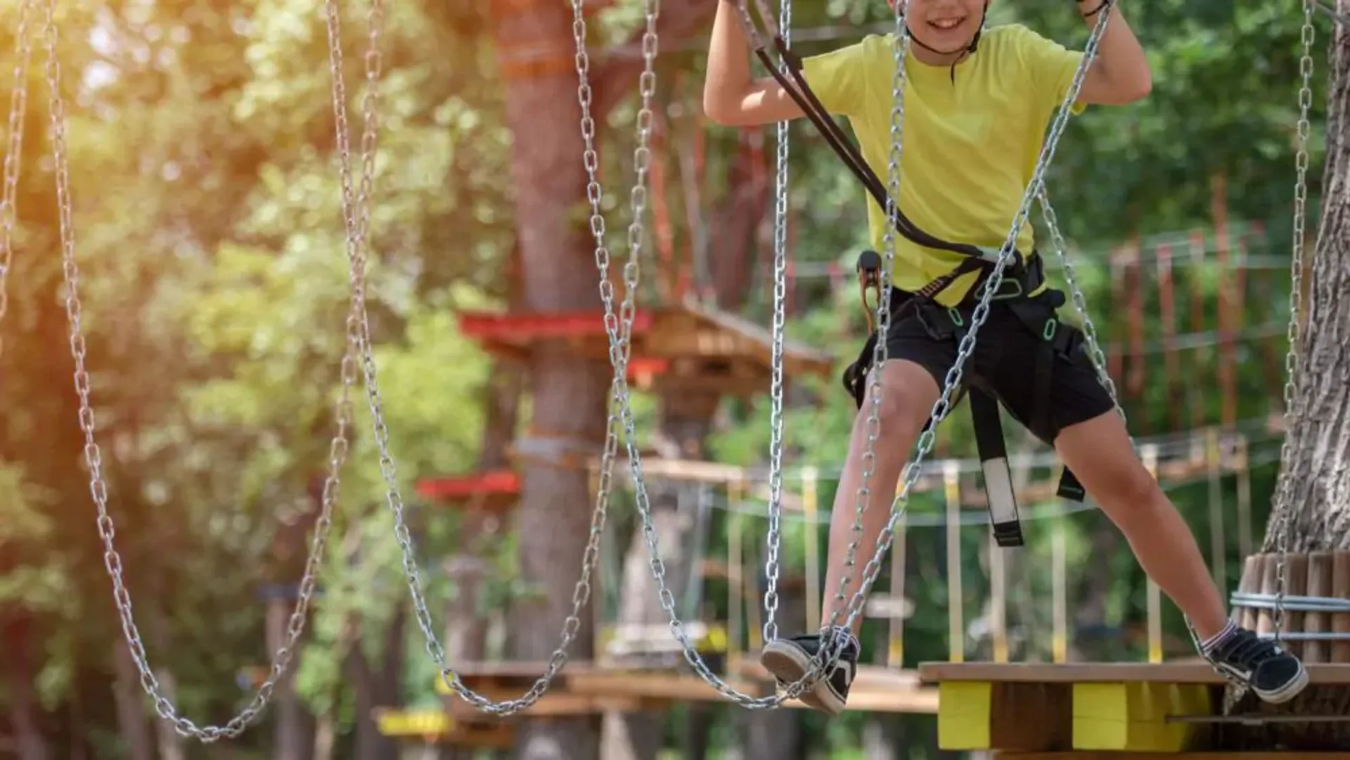 Ett barn klättrar och balanserar på en hög höjdsbana med säkerhetssele i en skogsmiljö. Kedjor och plattformar ingår i klätterbanan.