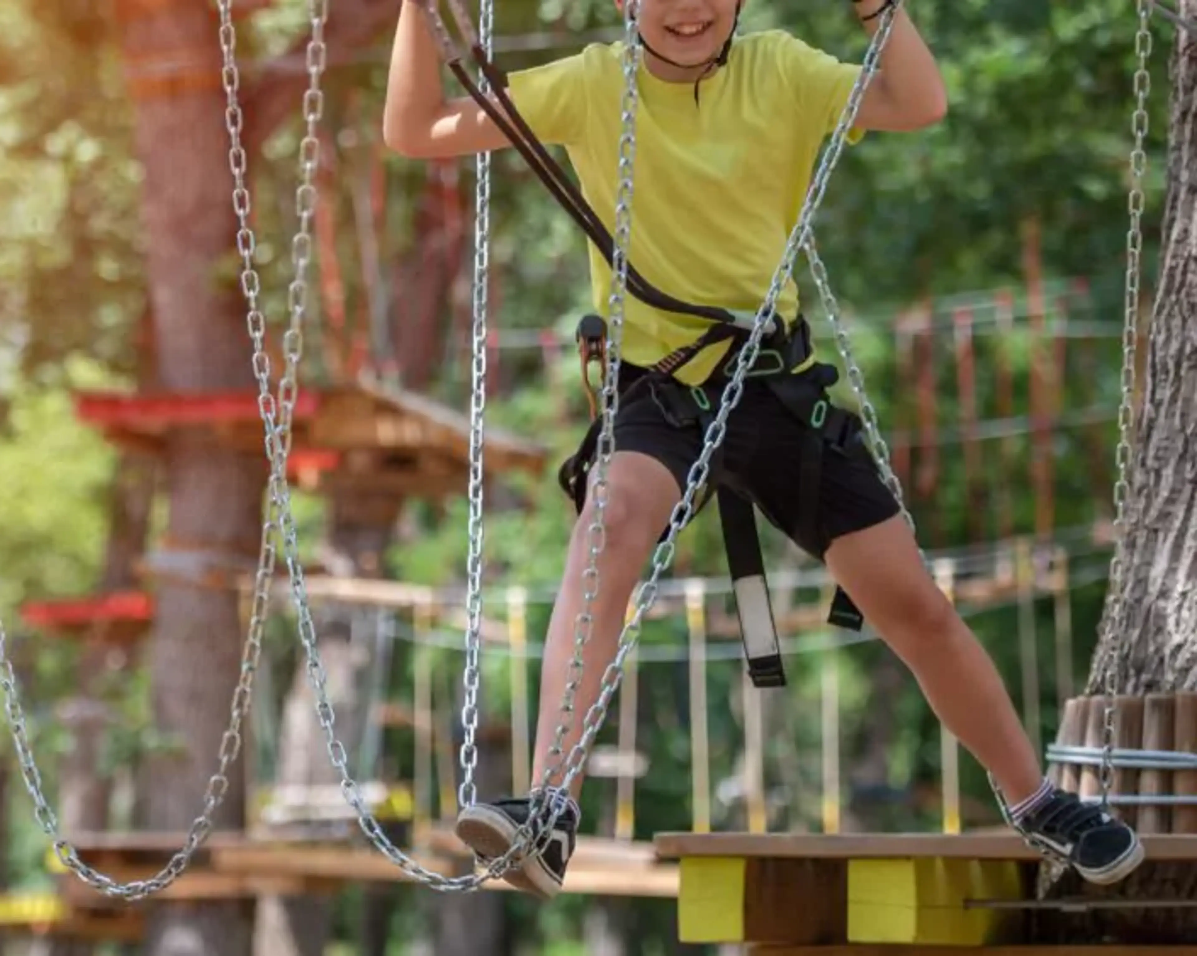 Ett barn klättrar och balanserar på en hög höjdsbana med säkerhetssele i en skogsmiljö. Kedjor och plattformar ingår i klätterbanan.