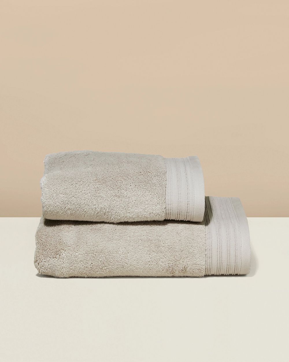 Ett set med två mjuka handdukar i beige med randig kantdetalj. Den större handduken ligger under den mindre, båda prydligt vikta.