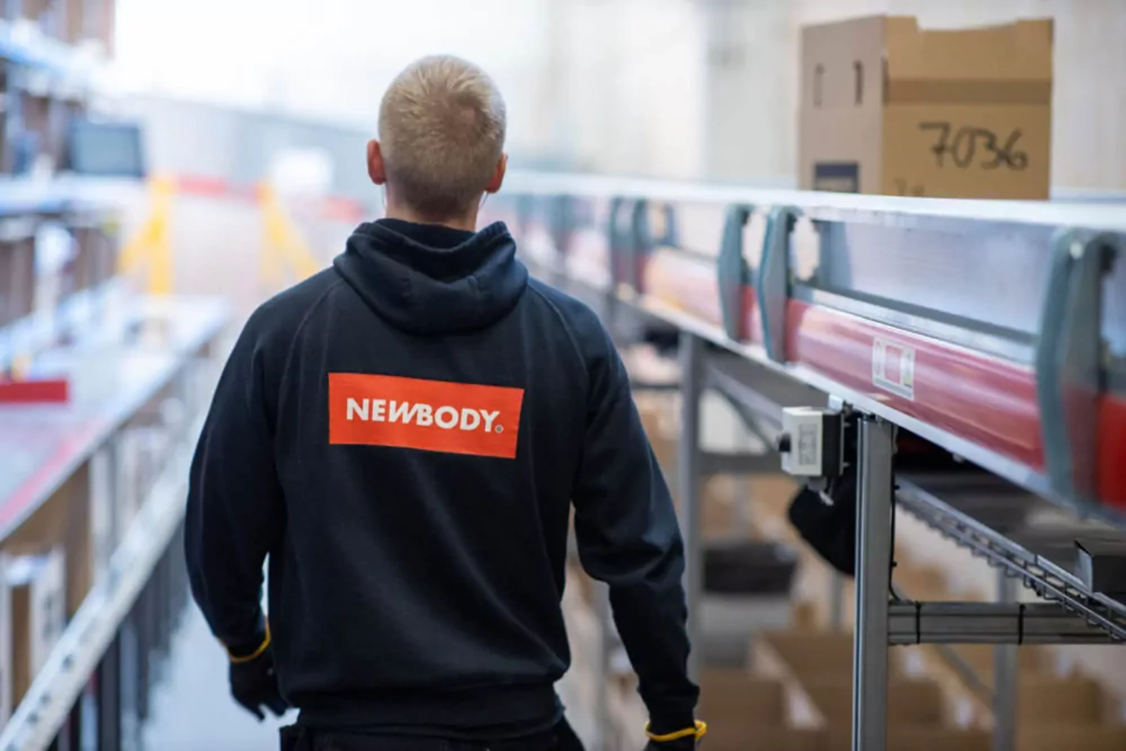 En person i en mörk huvtröja med en tydlig röd och vit logotyp på ryggen, som går längs en rad av hyllor och transportband i en lagerlokal.