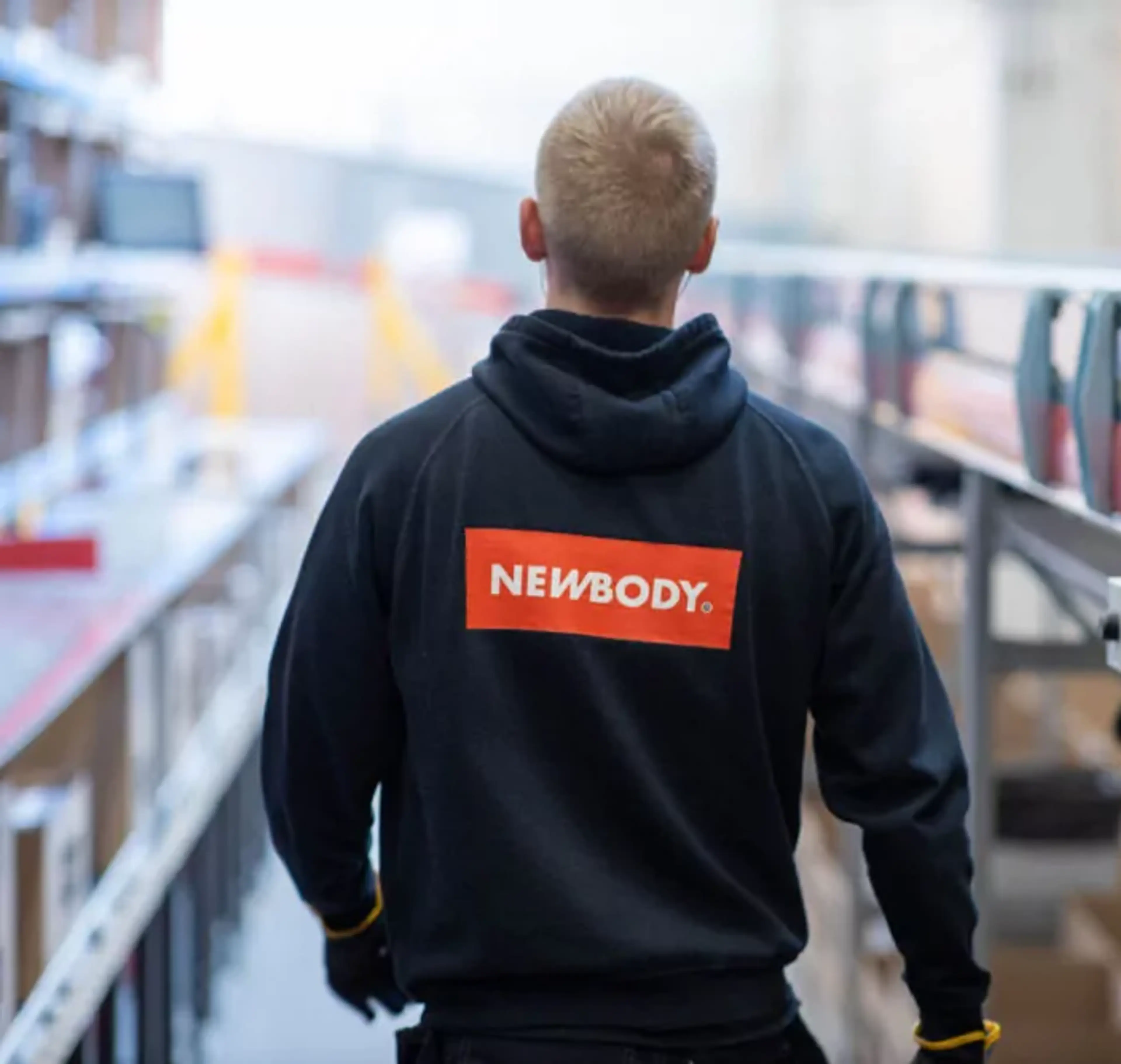 En person i en mörk huvtröja med en tydlig röd och vit logotyp på ryggen, som går längs en rad av hyllor och transportband i en lagerlokal.