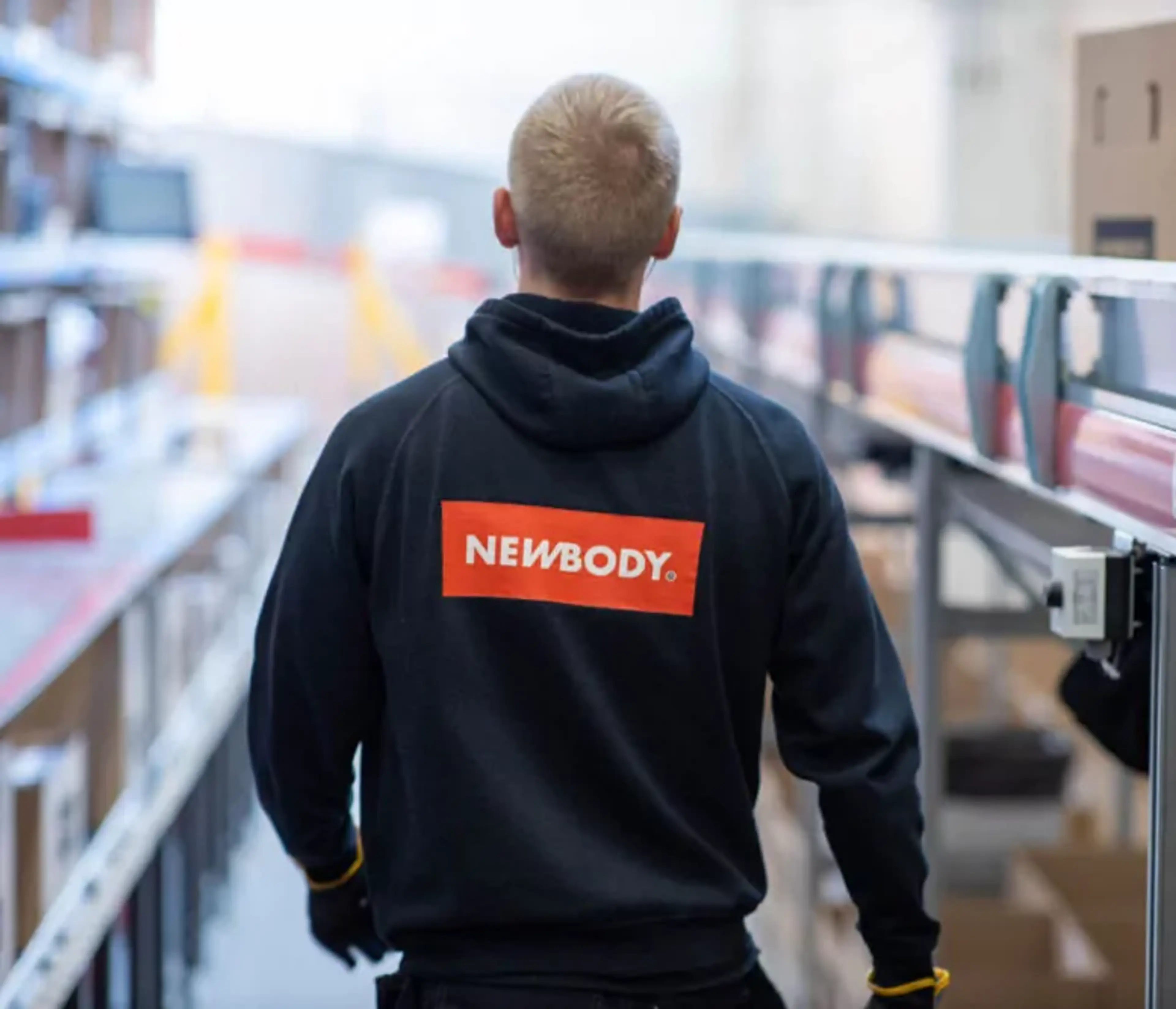 En person i en mörk huvtröja med en tydlig röd och vit logotyp på ryggen, som går längs en rad av hyllor och transportband i en lagerlokal.