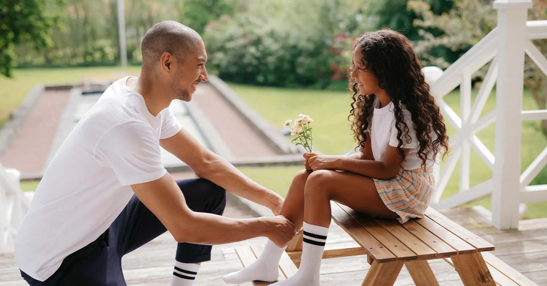 En vuxen man knyter vita sportstrumpor med svarta ränder på en flicka som håller små blommor, utomhus på en träbänk. Båda bär matchande strumpor och har på sig somriga kläder.