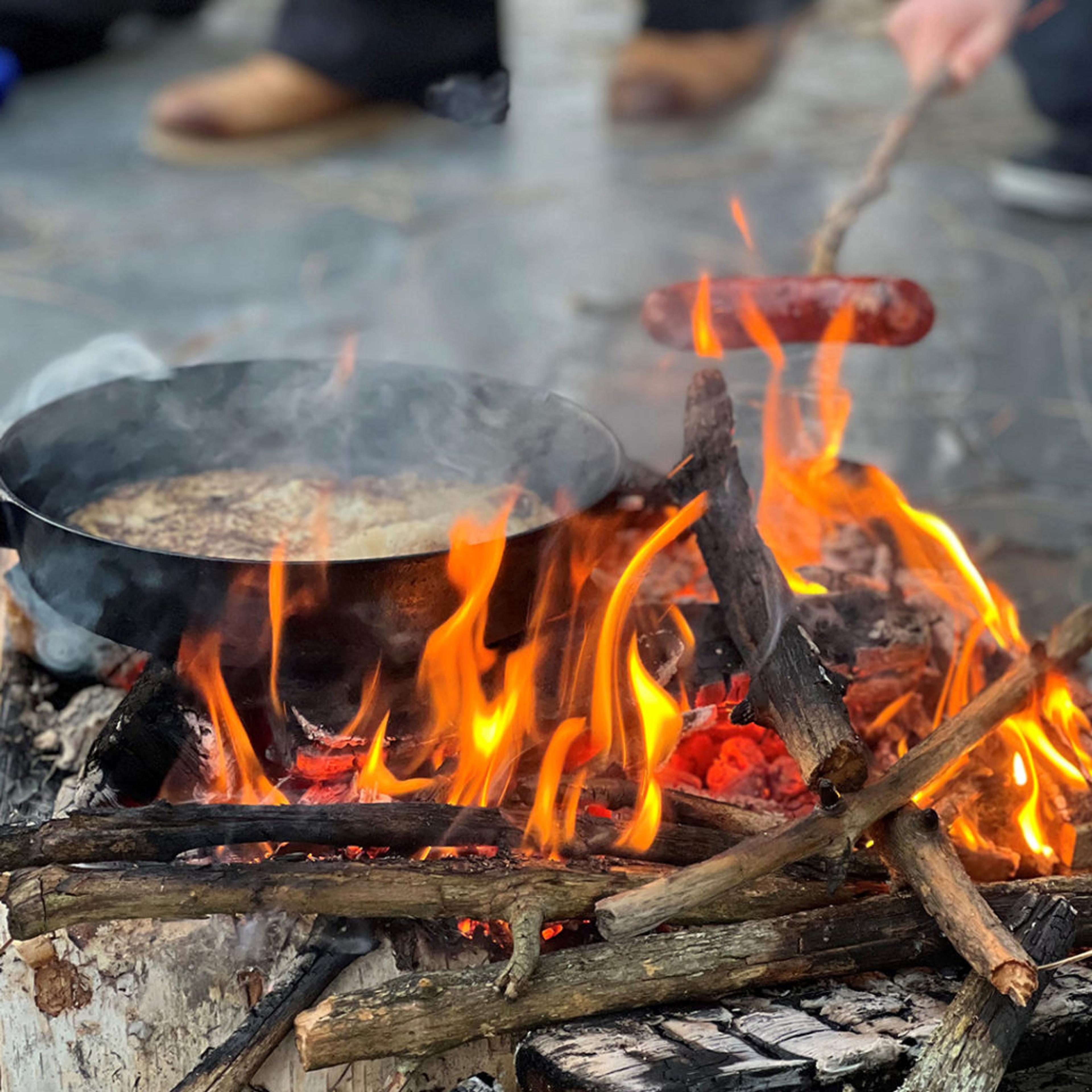 En stekpanna över öppen eld där något steks, och en korv som grillas på en pinne ovanför lågorna. Träbitar brinner och skapar en sprakande lägereld.