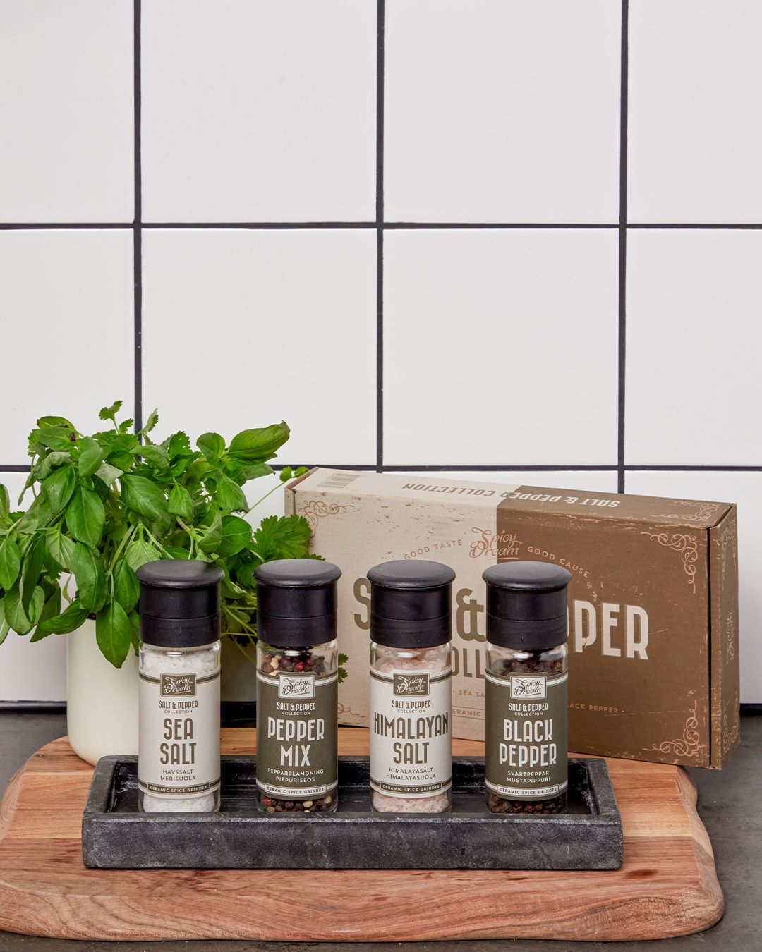 Fyra kryddburkar med olika salt- och peppersorter på en bricka, placerade på en skärbräda framför ett vitt kakel. En liten kruka med färska örter står bredvid.