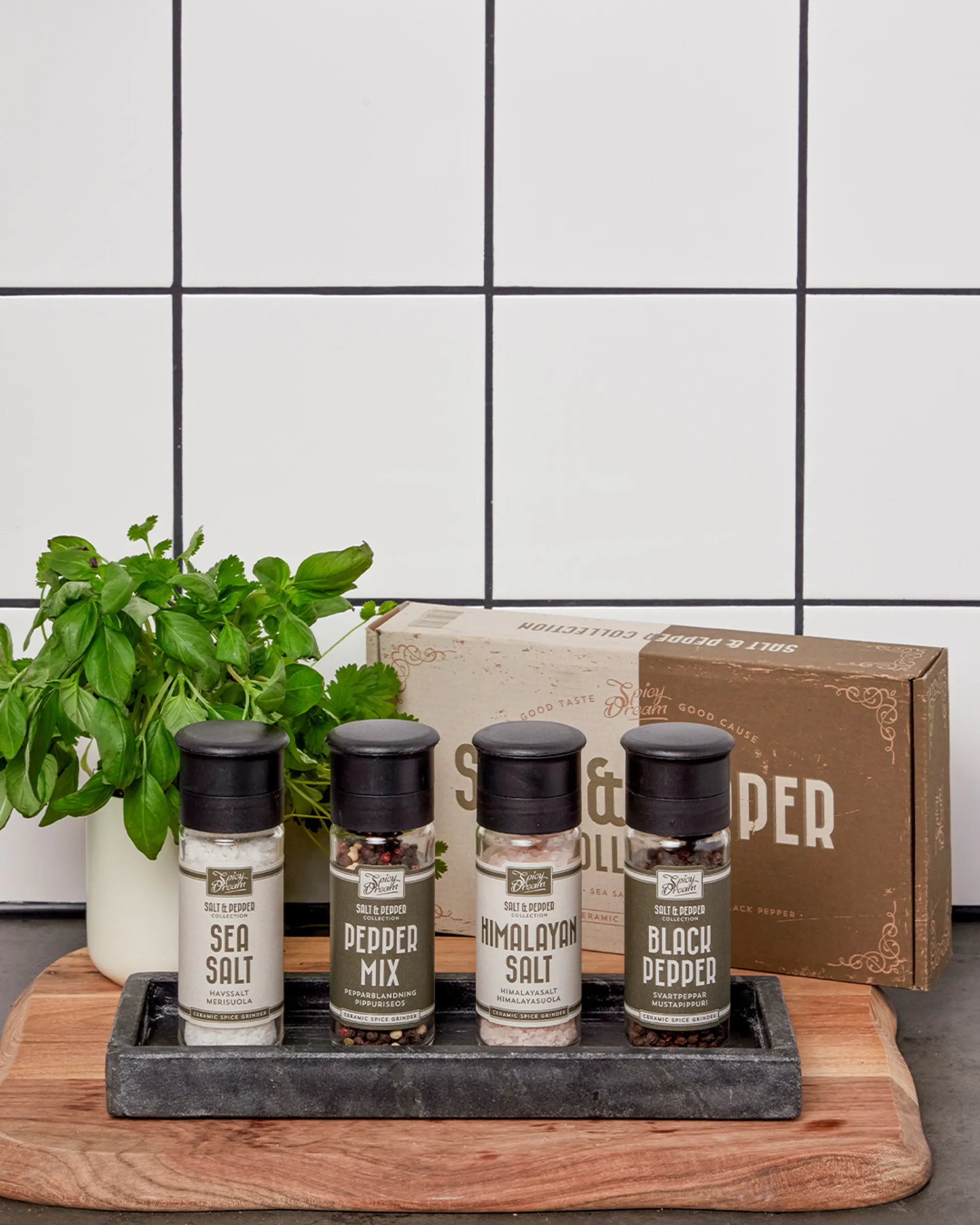 Fyra kryddburkar med olika salt- och peppersorter på en bricka, placerade på en skärbräda framför ett vitt kakel. En liten kruka med färska örter står bredvid.