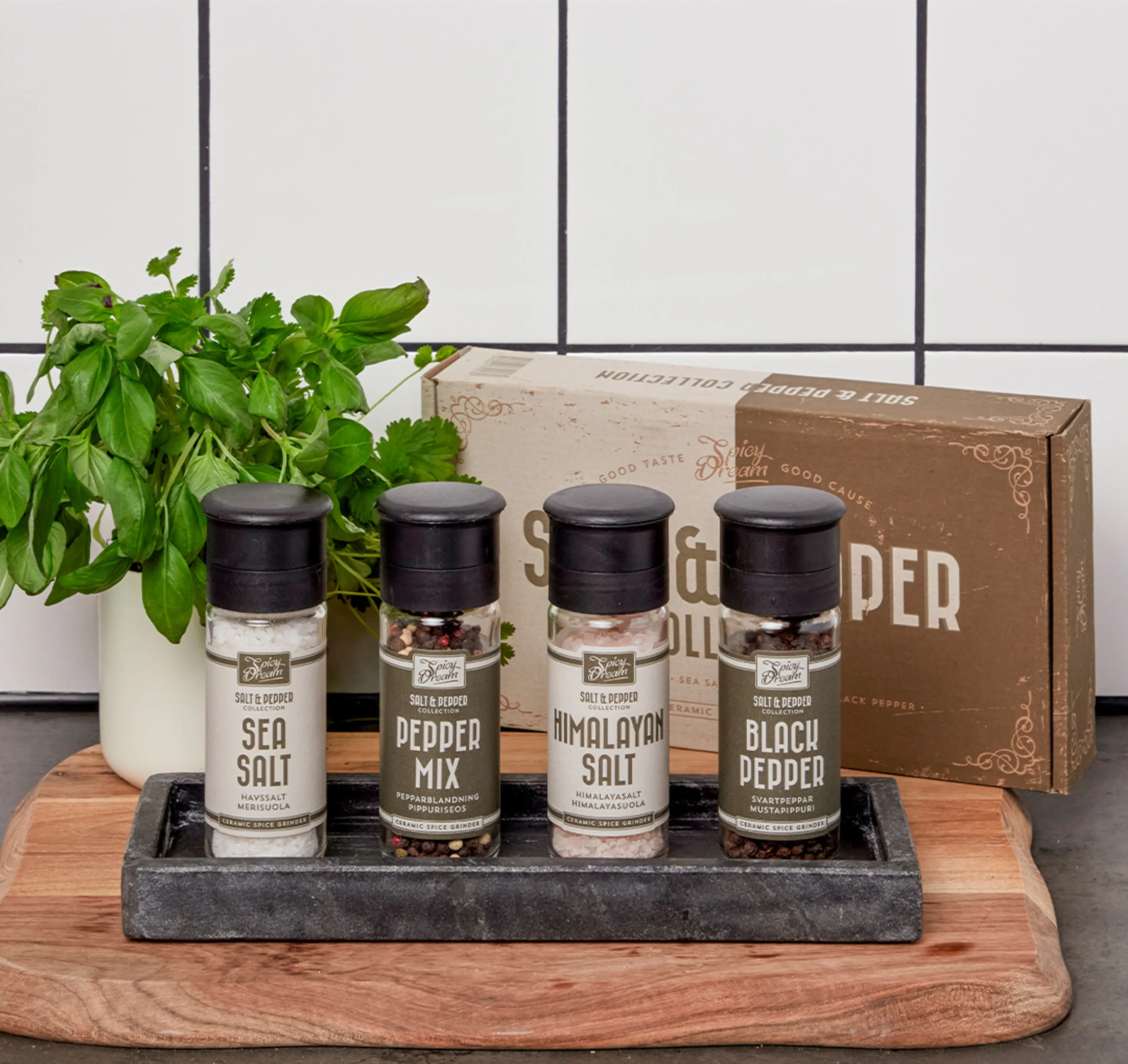 Fyra kryddburkar med olika salt- och peppersorter på en bricka, placerade på en skärbräda framför ett vitt kakel. En liten kruka med färska örter står bredvid.