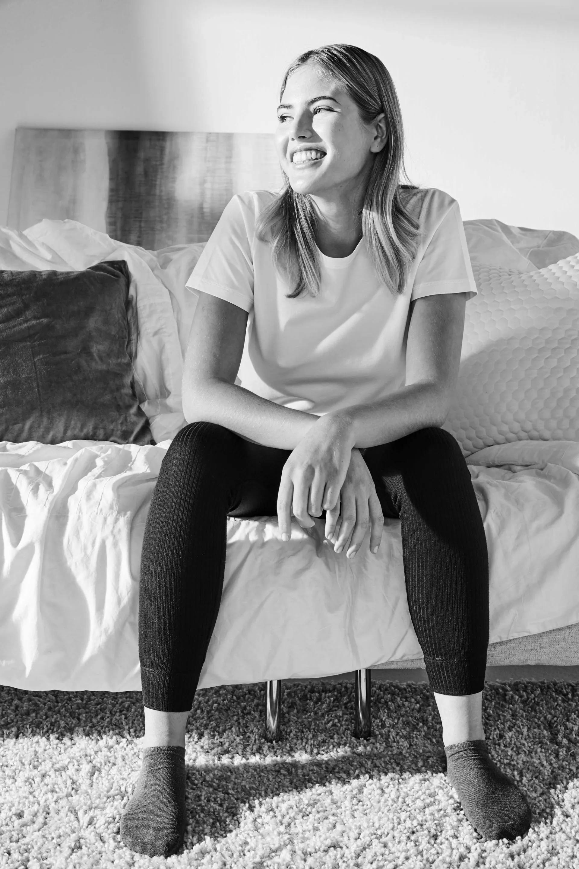 En person med vit t-shirt och svarta strumpbyxor sitter på en säng med ljusa sängkläder och kuddar. Golvet täcks av en mjuk, tjock matta.