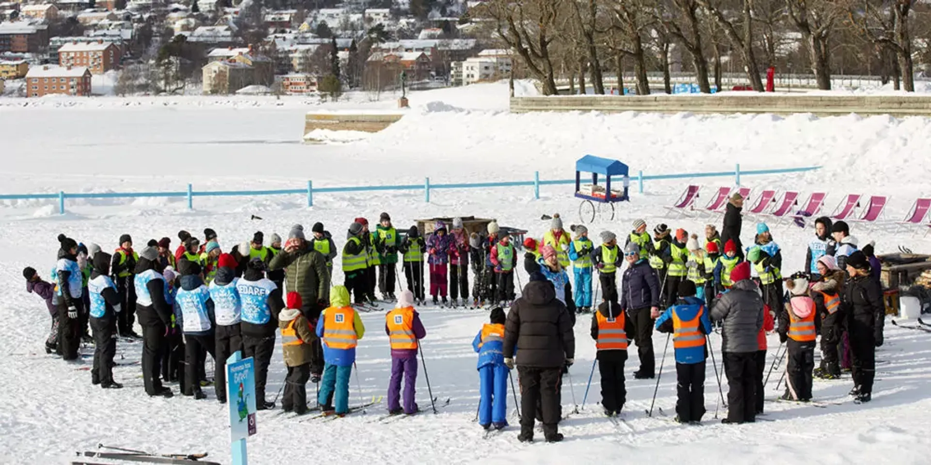 Barn och vuxna samlade i cirkel på en snötäckt yta, alla iklädda färgglada vinterkläder och säkerhetsvästar. Området är en öppen vinterscen med trädrad och byggnader i bakgrunden.