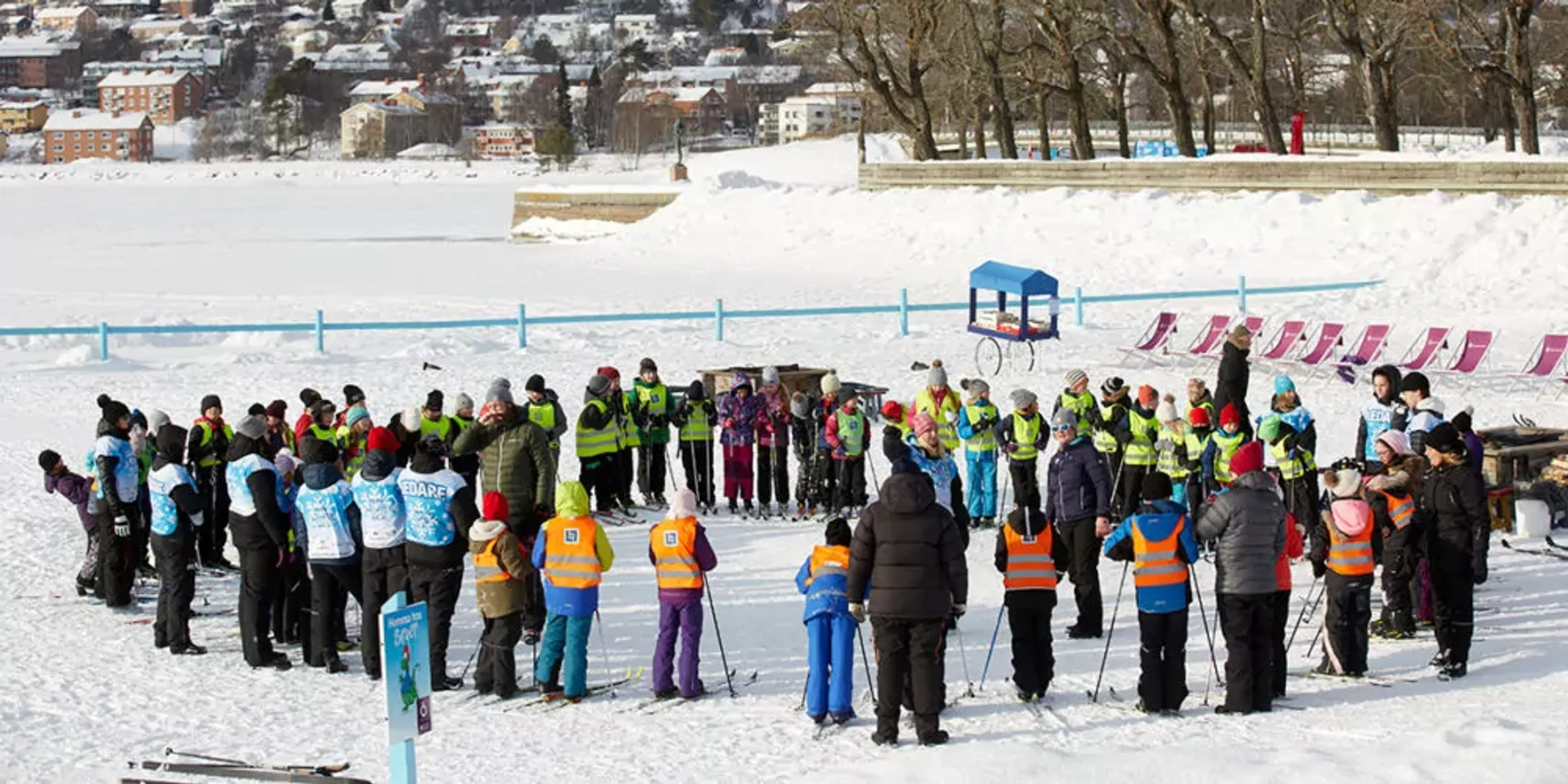 Barn och vuxna samlade i cirkel på en snötäckt yta, alla iklädda färgglada vinterkläder och säkerhetsvästar. Området är en öppen vinterscen med trädrad och byggnader i bakgrunden.