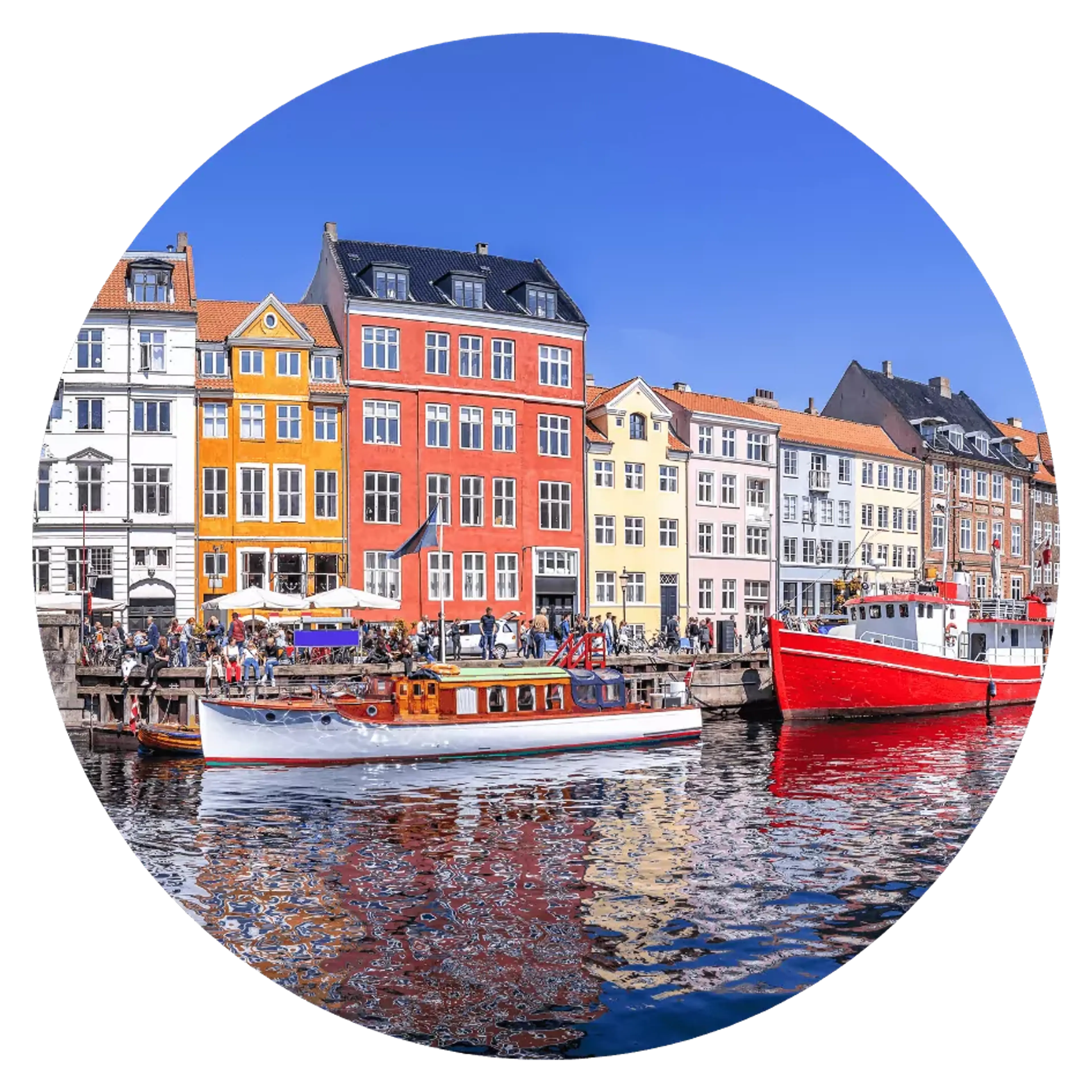 Färgglada byggnader längs med en kaj i en hamnstad med två fritidsbåtar i vattnet. Många människor samlas vid vattnet under en klarblå himmel.