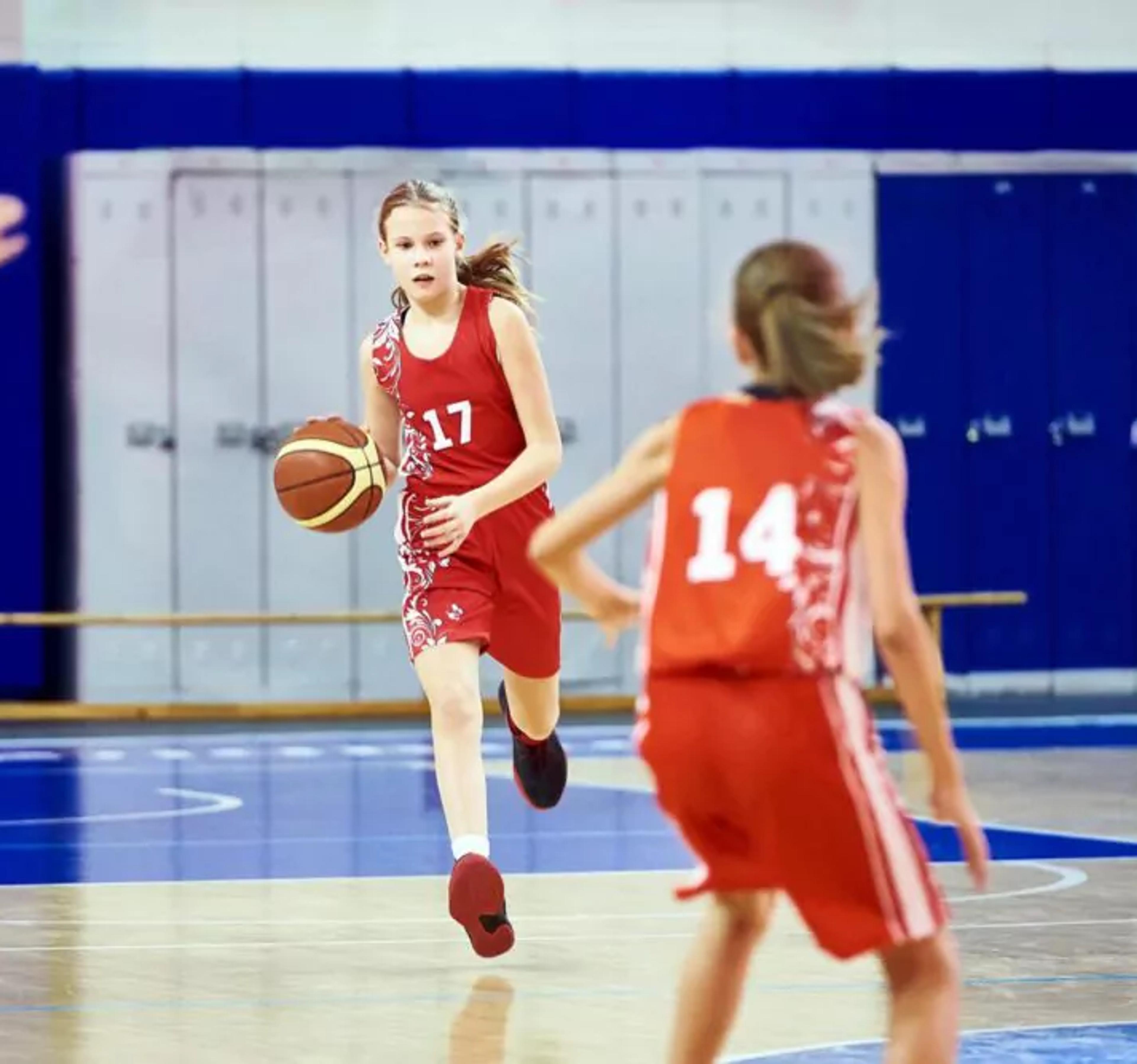 Tre unga spelare i röda matchställ spelar basket i en inomhushall med blå golv och vita linjer. En av dem dribblar bollen medan de andra rör sig på planen.