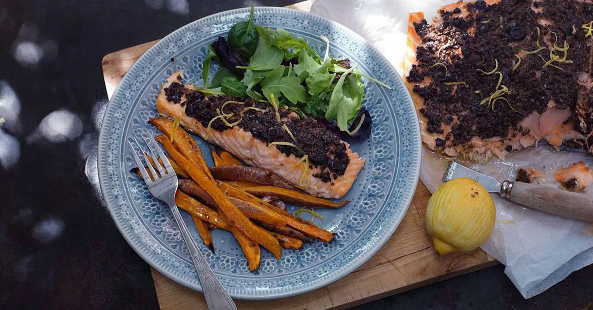 En portion ugnsbakad lax med tapenade, sötpotatisstavarna och en fräsch grönsallad på en blåmönstrad tallrik, bredvid ligger en bit lax med tapenade och en citron.