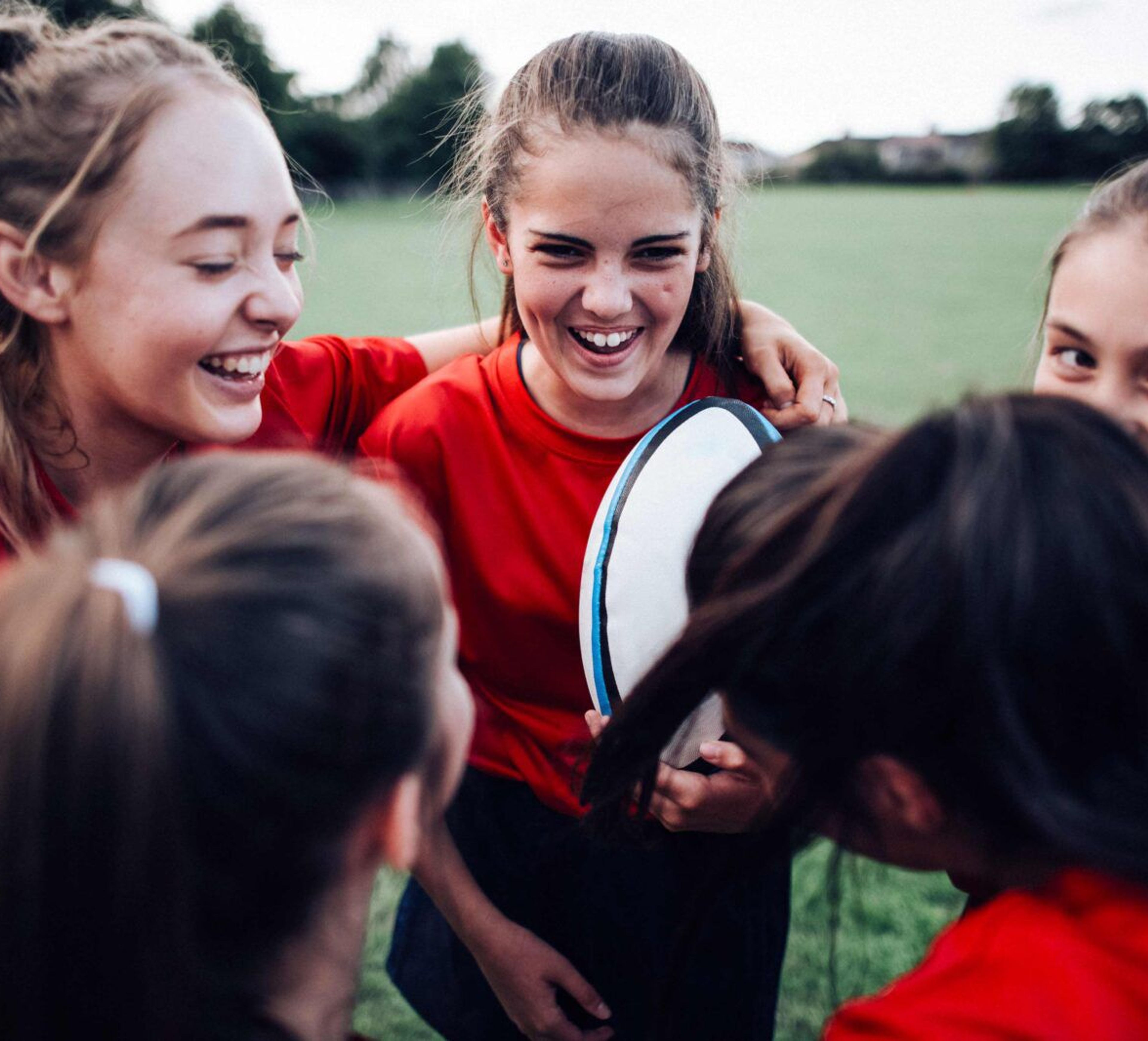 Fem flickor i röda tröjor står tätt tillsammans på en gräsplan, en av dem håller en rugbyboll. De visar glädje och samhörighet i stunden.