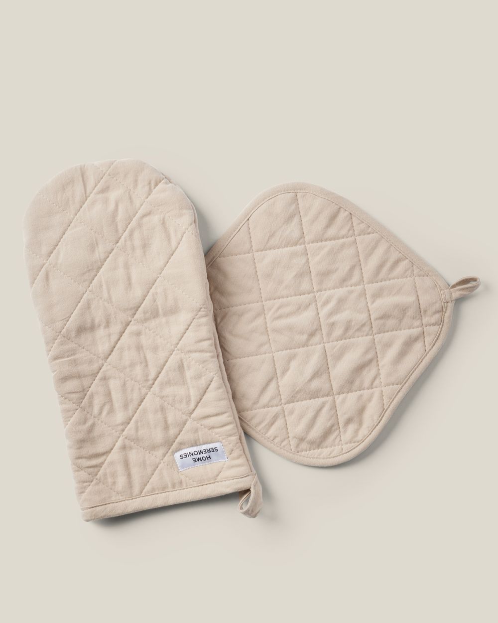 Ett set med en beige ugnsvante och en matchande grytlapp med quiltad design och öglor för upphängning. Mjuka och praktiska för köksbruk.