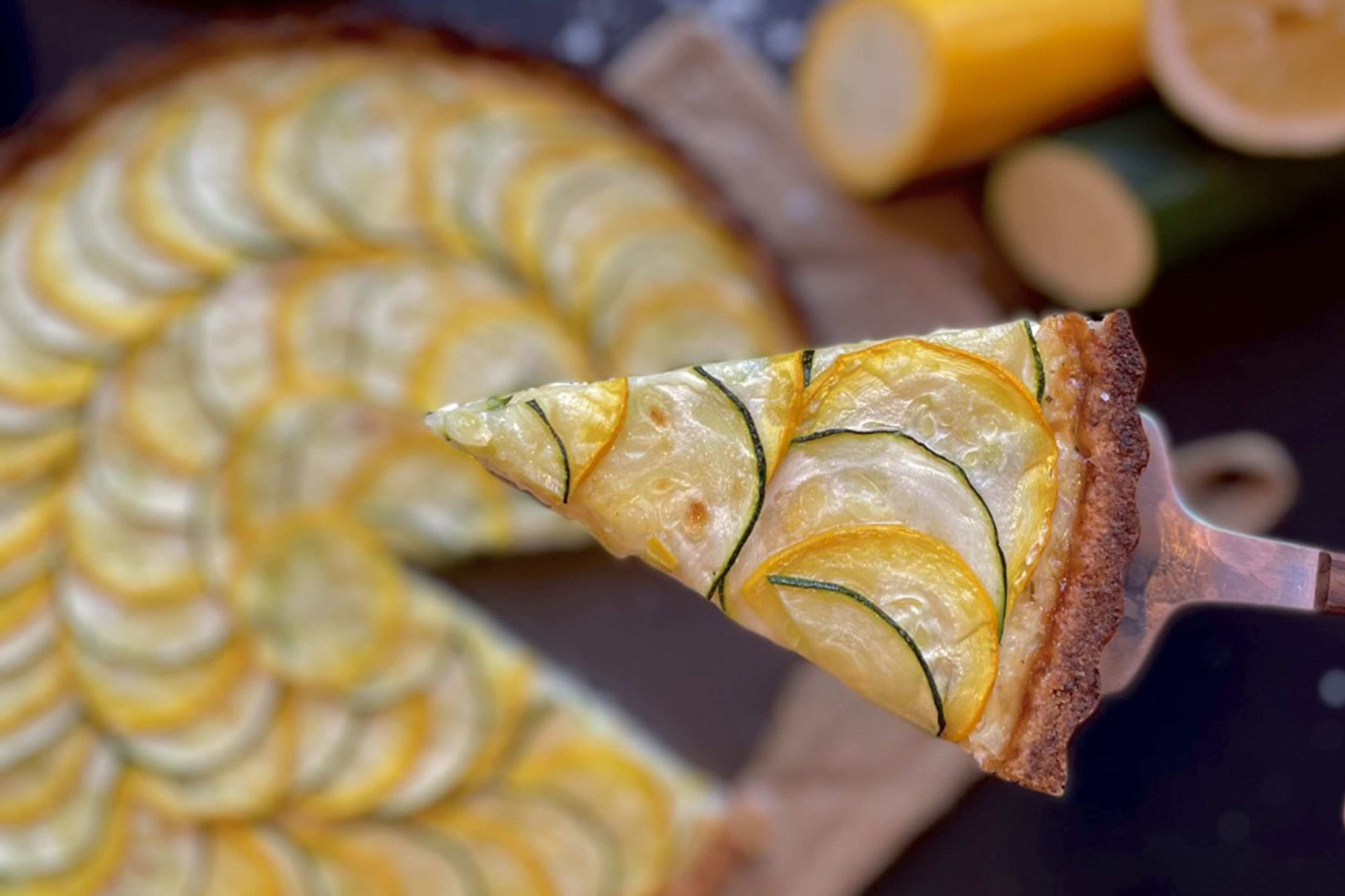 En skiva paj täckt med tunt skivade gula och gröna zucchiniskivor, fint upplagd i ett mönster. Pajskalet är gyllenbrunt och har en krispig kant.