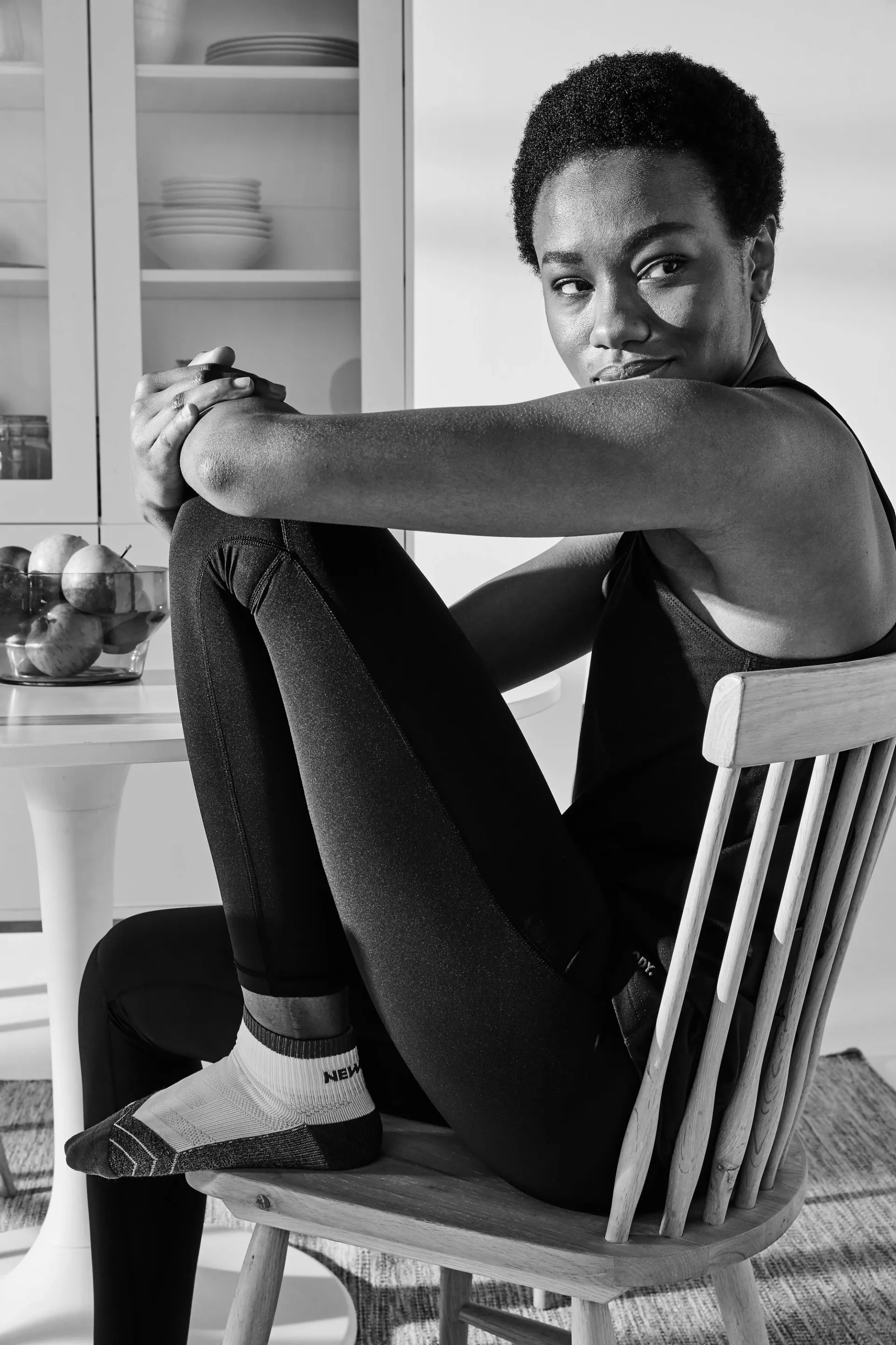 En person sitter på en trästol med knäna uppdragna och håller om ena armen, klädd i svart träningskläder och sportiga strumpor. I bakgrunden syns ett vitt köksskåp med tallrikar och en skål med ä