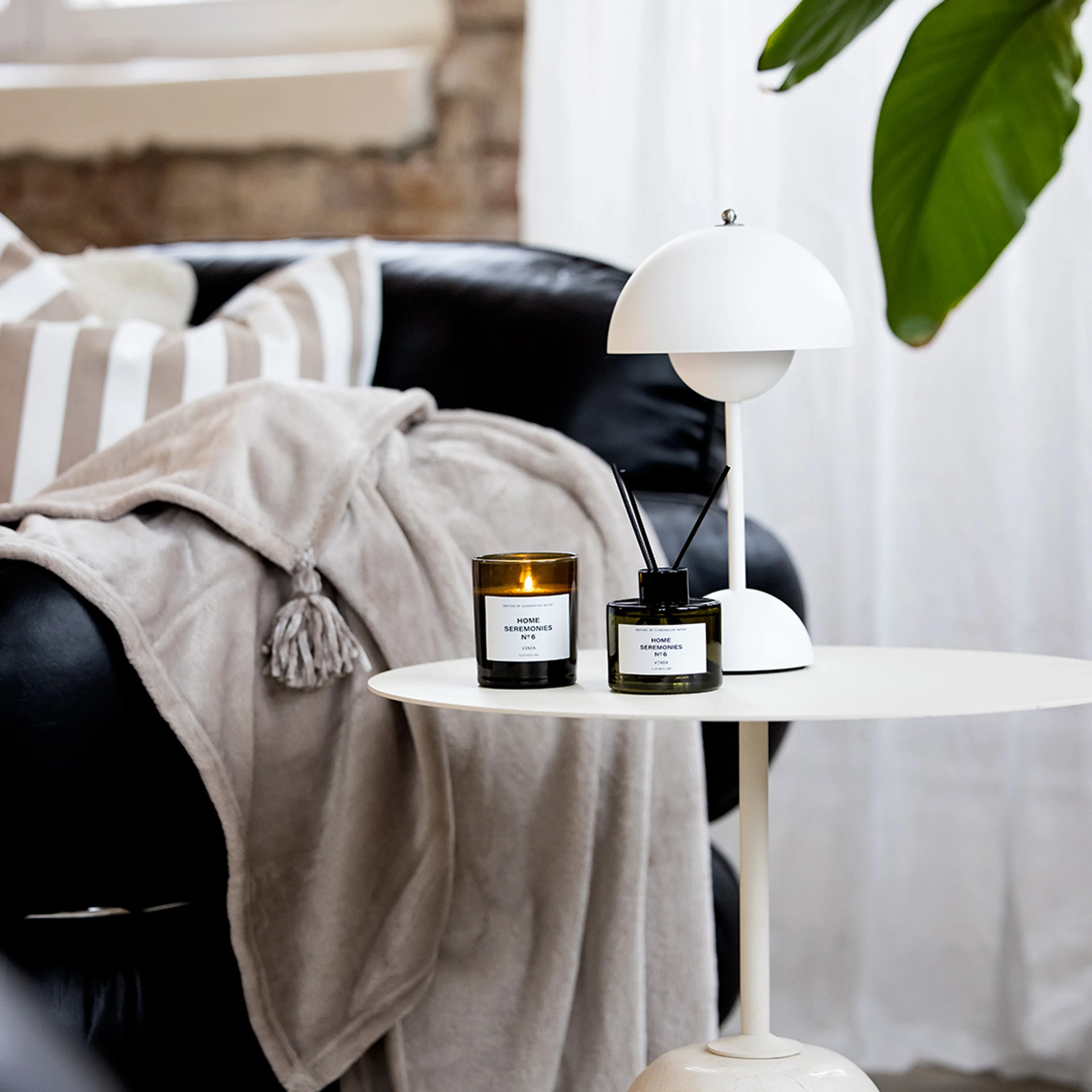 Ett doftljus och en rumsdoft med pinnar i mörkglas på ett runt vitt bord bredvid en vit bordslampa. I bakgrunden syns en svart fåtölj med en beige mjuk filt och en randig kudde.