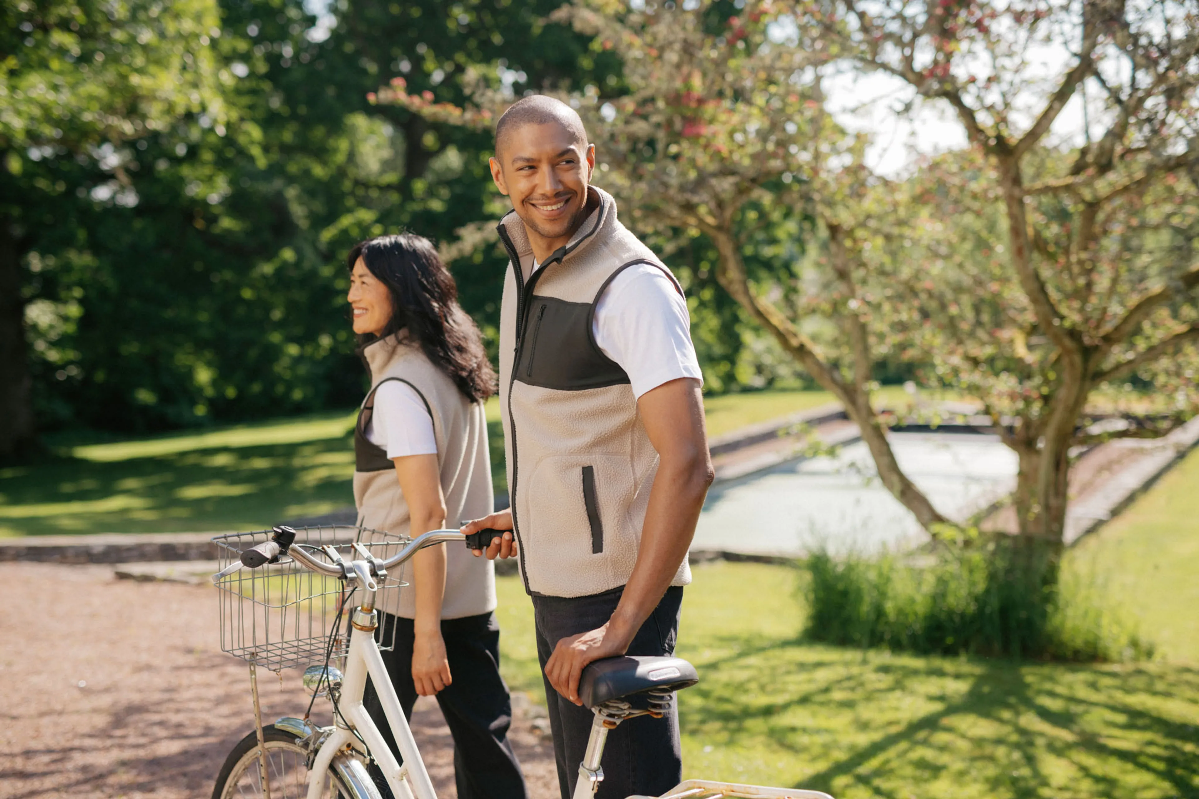 Två personer bär beige fleecevästar med svarta detaljer, den ena håller i en vit cykel. De står i en solig parkmiljö med gröna träd och gräsmatta i bakgrunden.