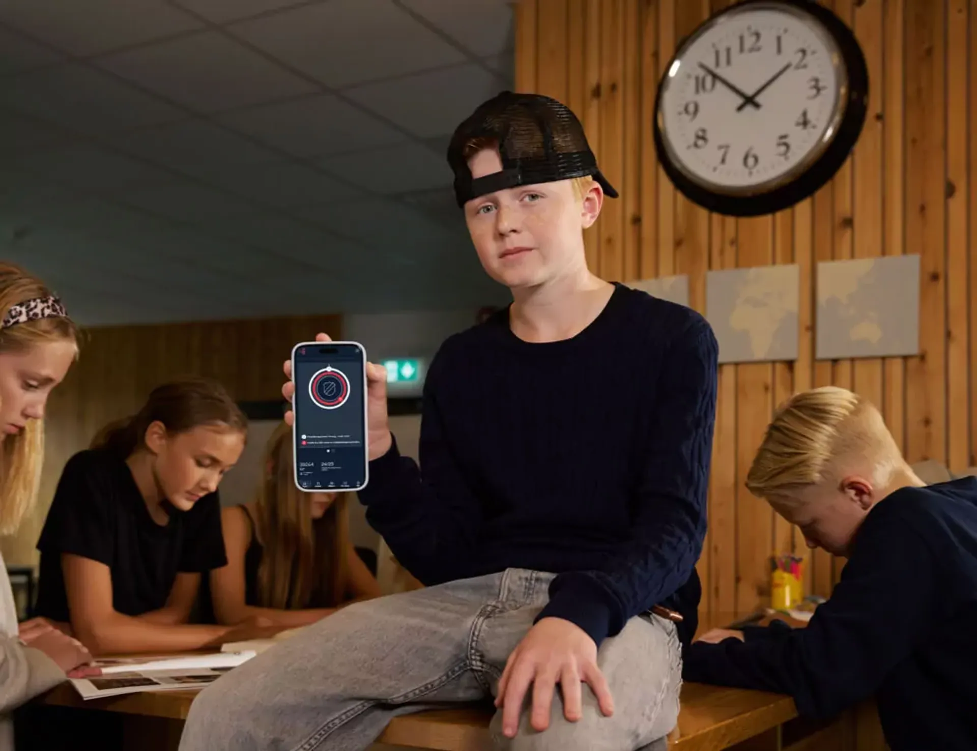 En grupp ungdomar sitter runt ett bord och studerar något, medan en kille med keps bak och mörk tröja visar en mobilskärm med en app. En rund väggklocka och världskartor syns på den träpanelklädda väggen.